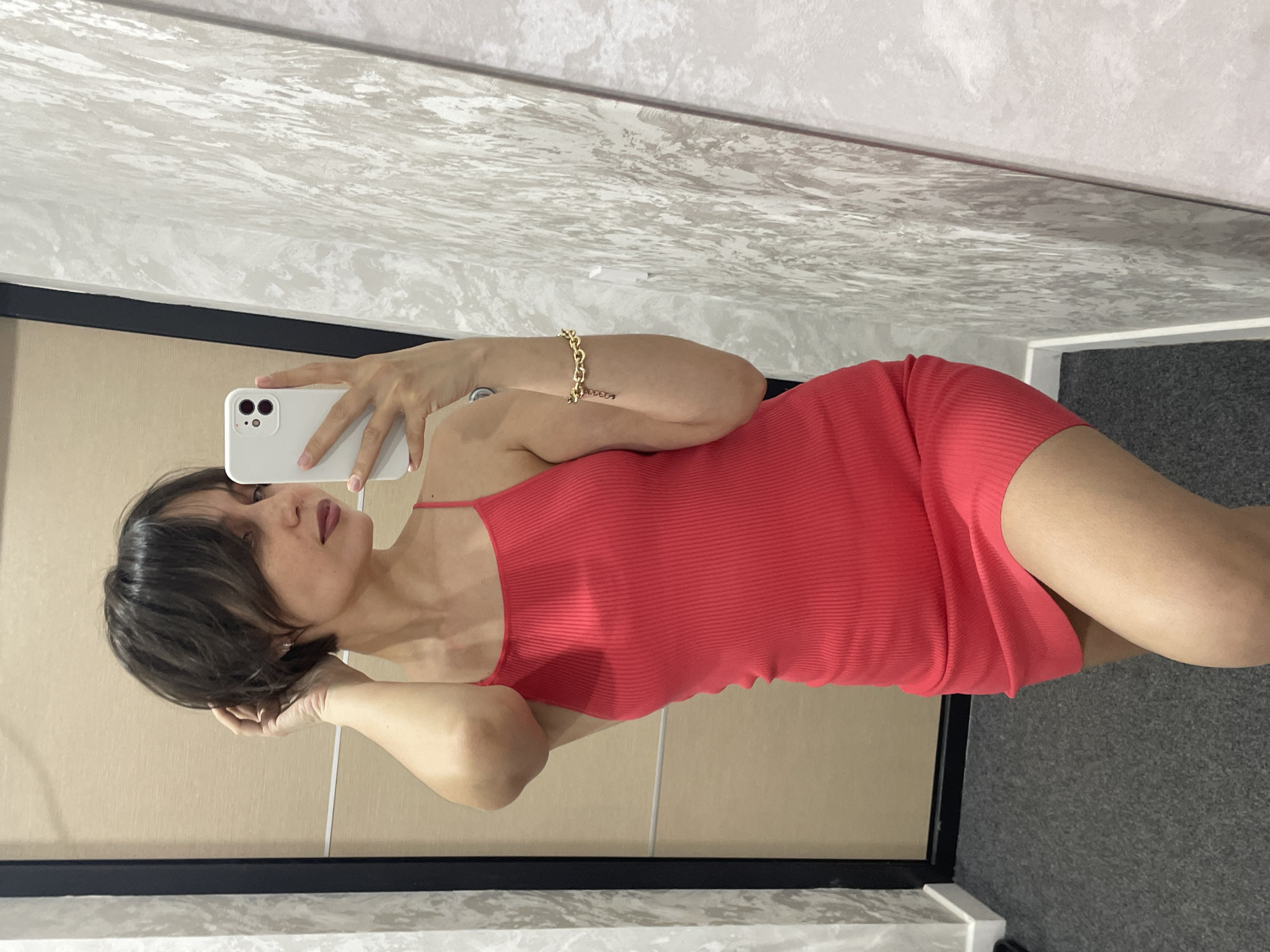 A woman is taking a selfie in what appears to be a dressing room. She is wearing a red, ribbed dress. She is holding a white smartphone. She has short, dark hair. She is wearing a gold bracelet on her left wrist. The background includes walls and flooring of different colors. The location is not possible to determine from the image.