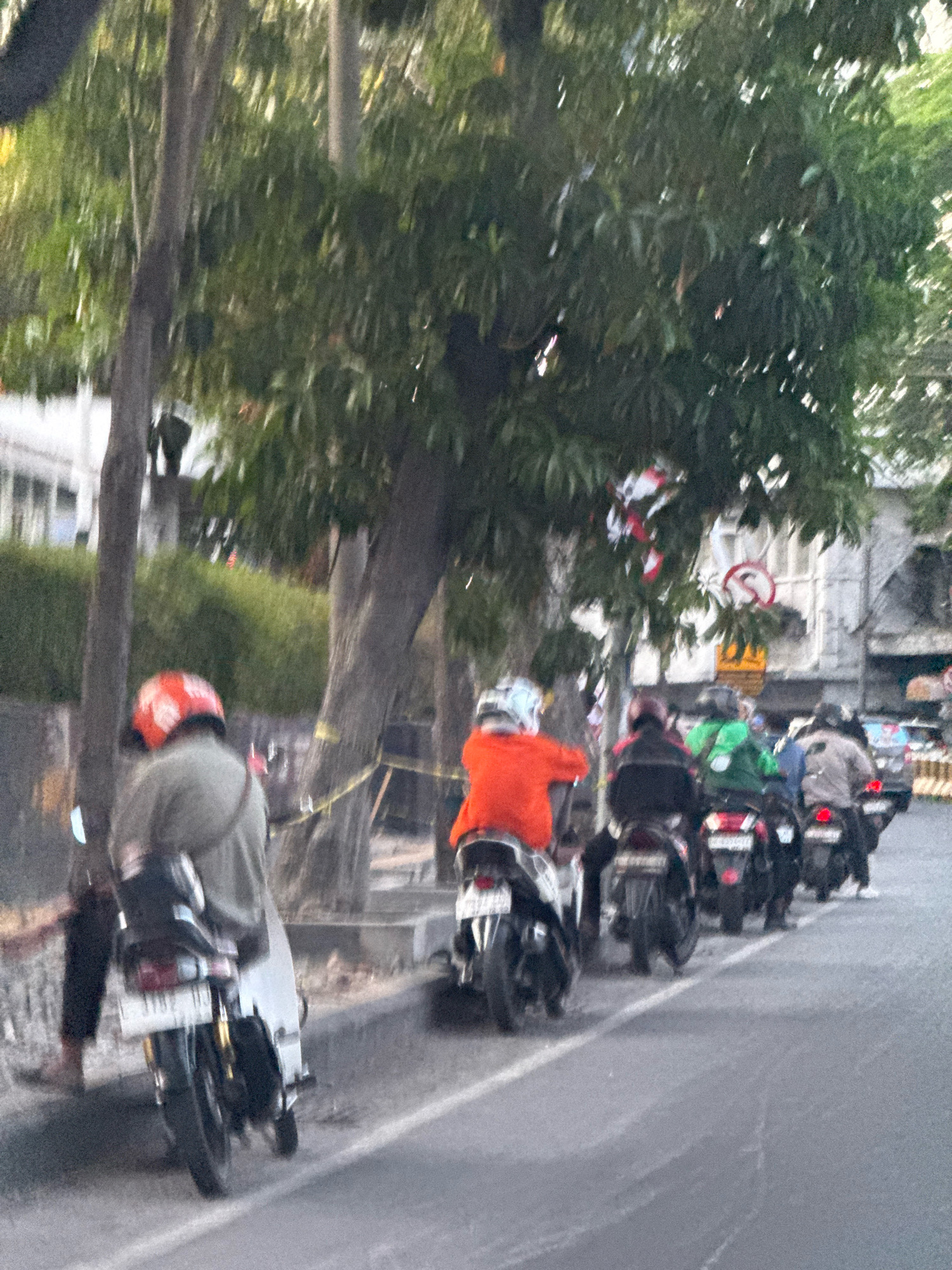 A group of motorcyclists are seen riding in a single file line on a street in Surabaya, Indonesia. The motorcyclists are wearing helmets and riding in the same direction. The street is lined with trees and buildings, and there is a sidewalk on the left side of the road. The image appears to be taken during the daytime, as there is natural light visible.
