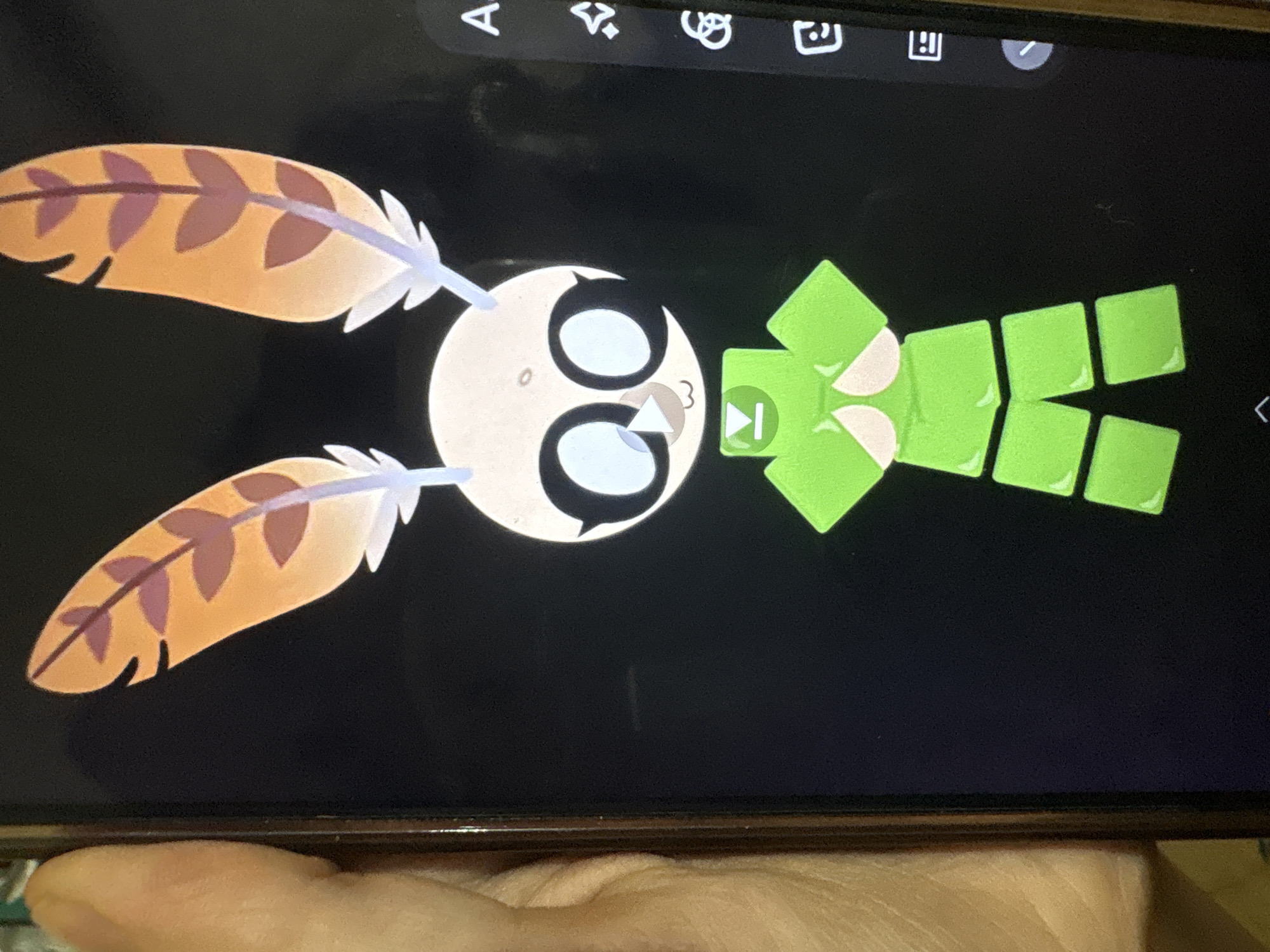 The image displays a digital illustration of a cartoon character on a smartphone screen. The character has a pale, round head with large, black-rimmed eyes with light blue irises. It has two light purple, feather-like antennae on top of its head, each adorned with brown, leaf-like shapes. The character's mouth is small and appears to be smiling. Its body is composed of green, block-like shapes. A small white triangle is positioned below the character's mouth.
The smartphone screen is dark, with a black background. On the right side of the screen, there is a vertical toolbar with several icons, including an "A", a starburst, a circular arrow symbol, a speech bubble icon, and a horizontally divided rectangle icon. A faint reflection is visible on the screen.
A person's left hand and thumb, with visible skin texture and fingernails, are partially visible on the left side of the image, holding the smartphone. The background behind the phone is out of focus and appears to be a textured surface, possibly fabric or wood. There is no indication of the location being in Ashvem, India.