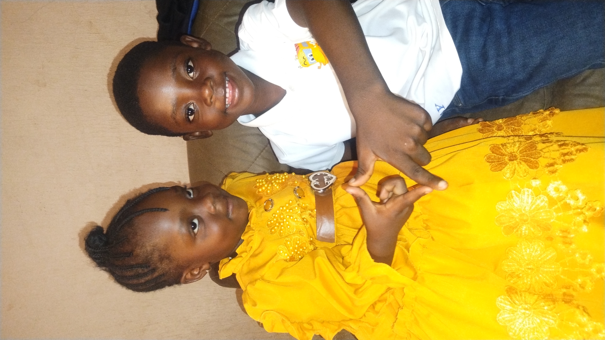Two young children, one boy and one girl, are lying on a couch in an indoor setting. The boy is lying on his back with his head tilted to the right and he is wearing a white t-shirt with a cartoon image and blue jeans. The girl is lying on her side in a bright yellow dress with a belt and flower embellishments. She has her hair styled in braided buns. Both children are looking at the camera, and the girl is making a finger gesture. The location is Jos, Nigeria. The background is a plain wall.