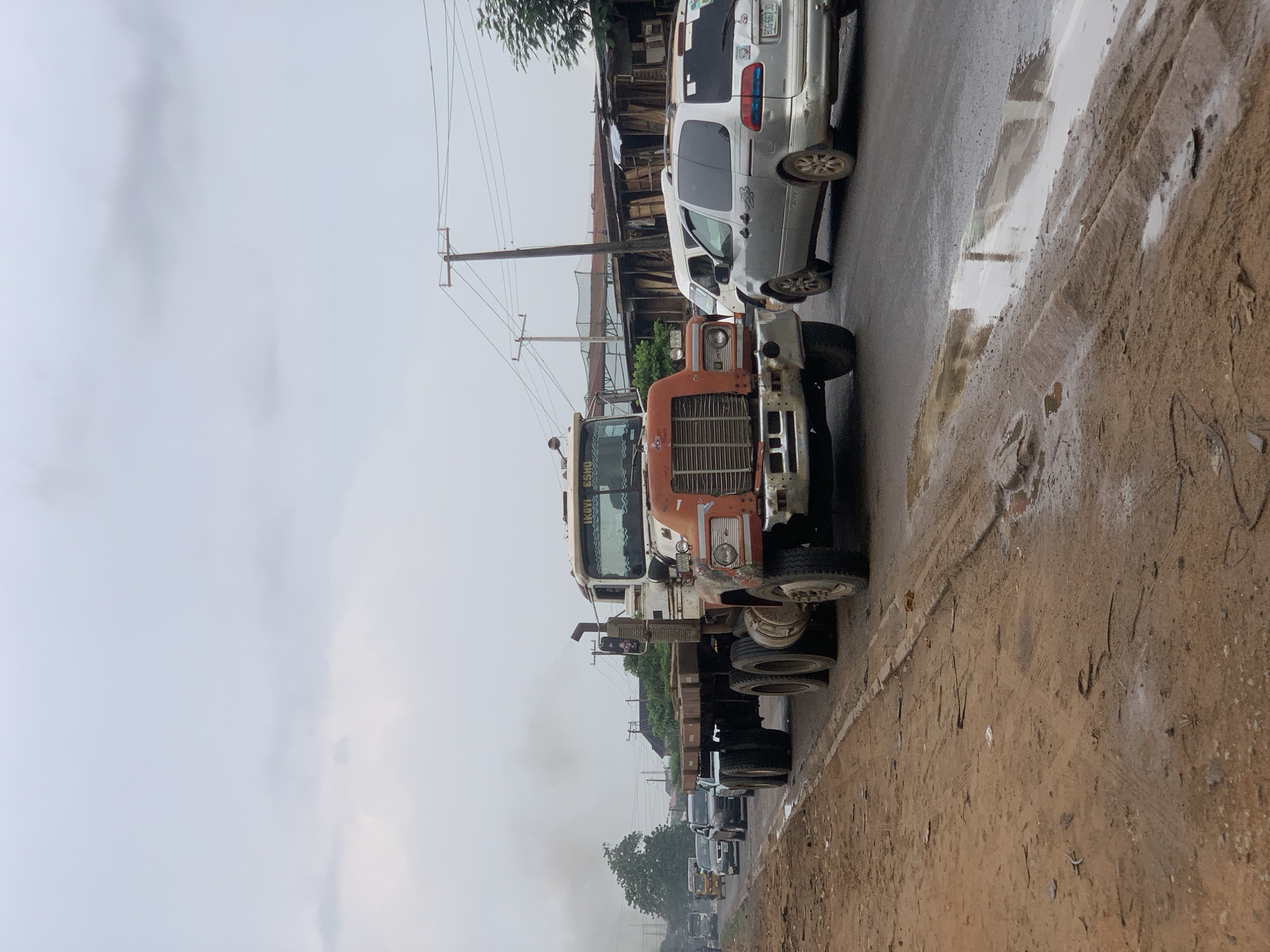 This image captures a busy roadside scene in Ikorodu, Nigeria, under an overcast, grey sky, suggesting either recent rain or an extremely cloudy day. The asphalt road is visibly wet with puddles reflecting the dull sky, and the unpaved shoulder is muddy and dirty.
In the foreground, a large, older model orange and white semi-truck or lorry, possibly a Mack truck, dominates the view, positioned on the wet road and facing towards the viewer. The truck shows signs of wear, with visible rust and grime on its front grille and bumper. Faint text, possibly "KOVI ESMAO," is discernible on its sun visor. To the right, the road's edge is a muddy, unpaved strip of earth with tire marks.
Behind the large truck, a silver minivan is parked on the road, partially obscured. Its license plate, "BGG 667 JD," is clearly visible. Further down the road, a queue of other vehicles, including more trucks and cars, extends into the hazy distance. A plume of smoke is rising in the background, possibly from one of the vehicles or nearby activity.
The scene includes several wooden utility poles carrying multiple overhead power lines, stretching along the roadside. In the background, various modest buildings, some appearing to be simple wooden or corrugated iron structures, are interspersed with scattered trees and greenery. The overall impression is that of an active, functional street environment in a semi-urban setting, marked by its robust traffic and basic infrastructure.