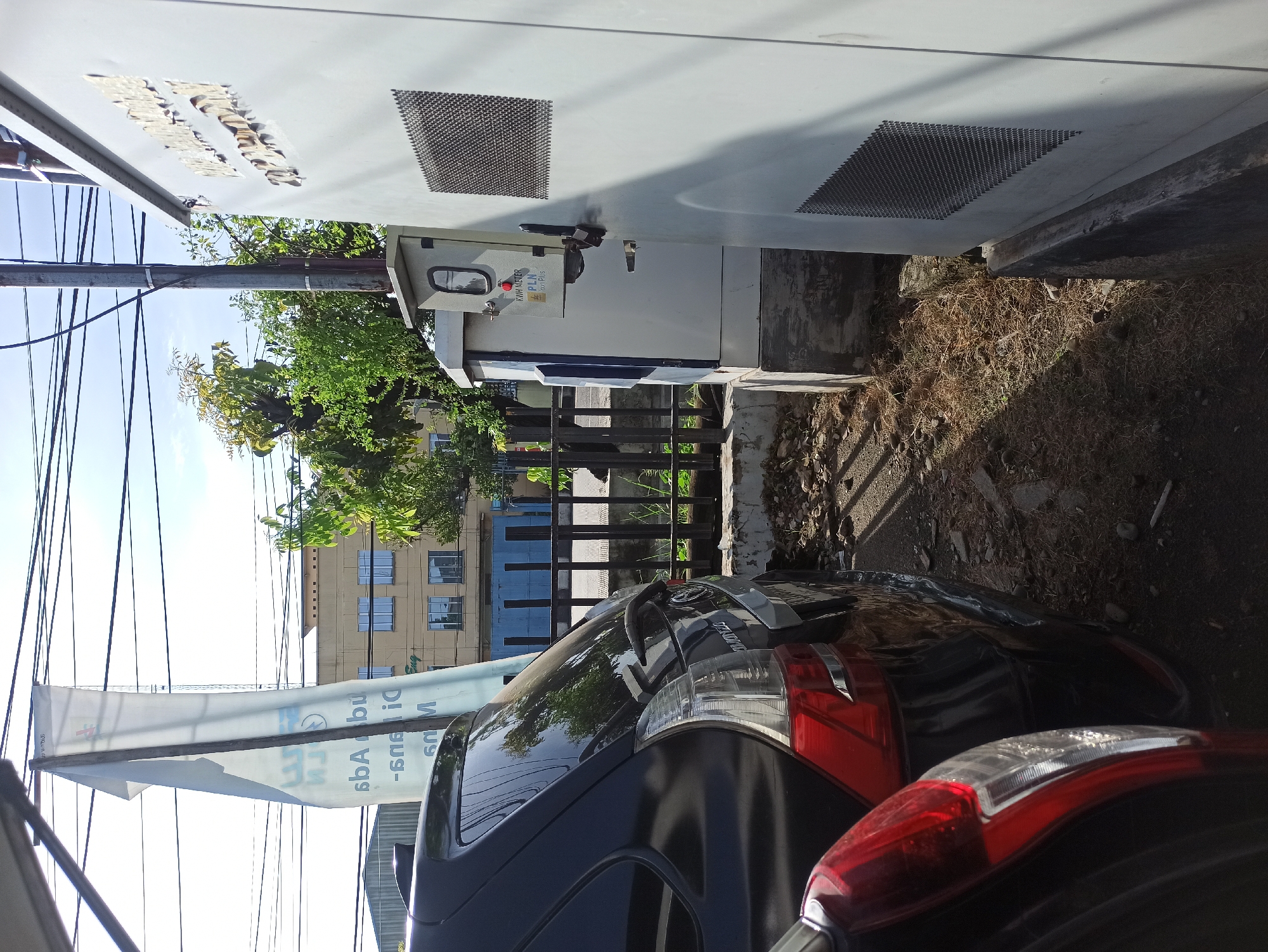 In the city of Medan, Indonesia, a black car is parked outdoors next to a white electrical utility box. The car is viewed from the rear, with its taillight and part of the bumper visible on the left side of the frame. The electrical box is tall and gray, with a locked door and a mesh vent on its front. A smaller, gray box with a window and a red button is attached to the larger utility box. A white banner with text in an Asian language is hanging from a pole behind the car, partially obscuring a building with multiple windows in the background. Trees with green leaves are visible behind the banner and electrical box, and power lines stretch across the sky. The ground is covered with dry grass and debris.