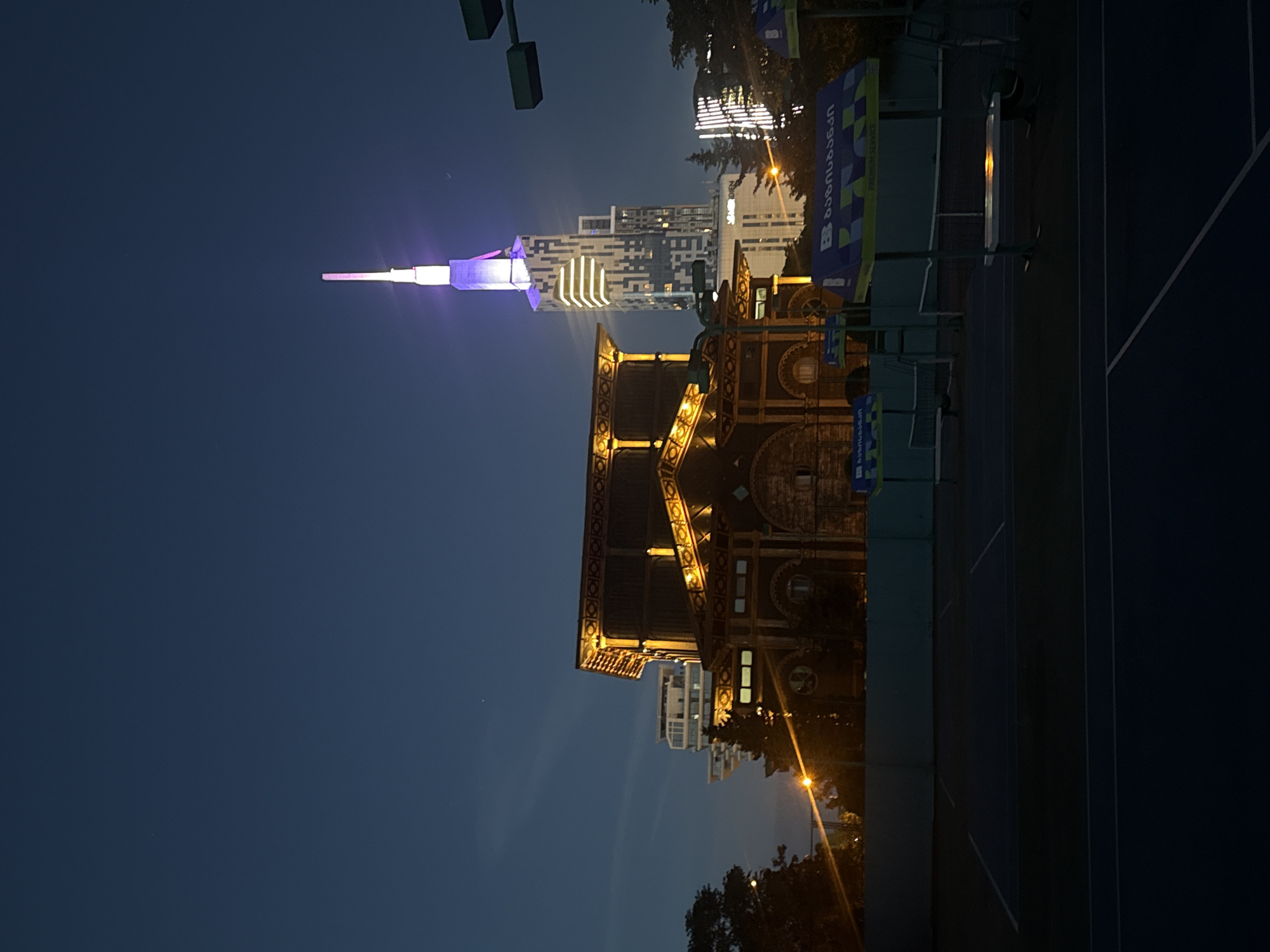 The image shows a night scene in Batumi, Georgia, featuring illuminated structures and urban elements. In the distance, a tall, modern building with a spire, illuminated with white and purple light, stands out against the dark sky. Nearer to the camera is an older, more ornate building with detailed architectural features, lit with warm yellow lights. A tennis court with equipment lines the right side of the shot, along with the signs and equipment next to it. Trees and street lights are also visible.