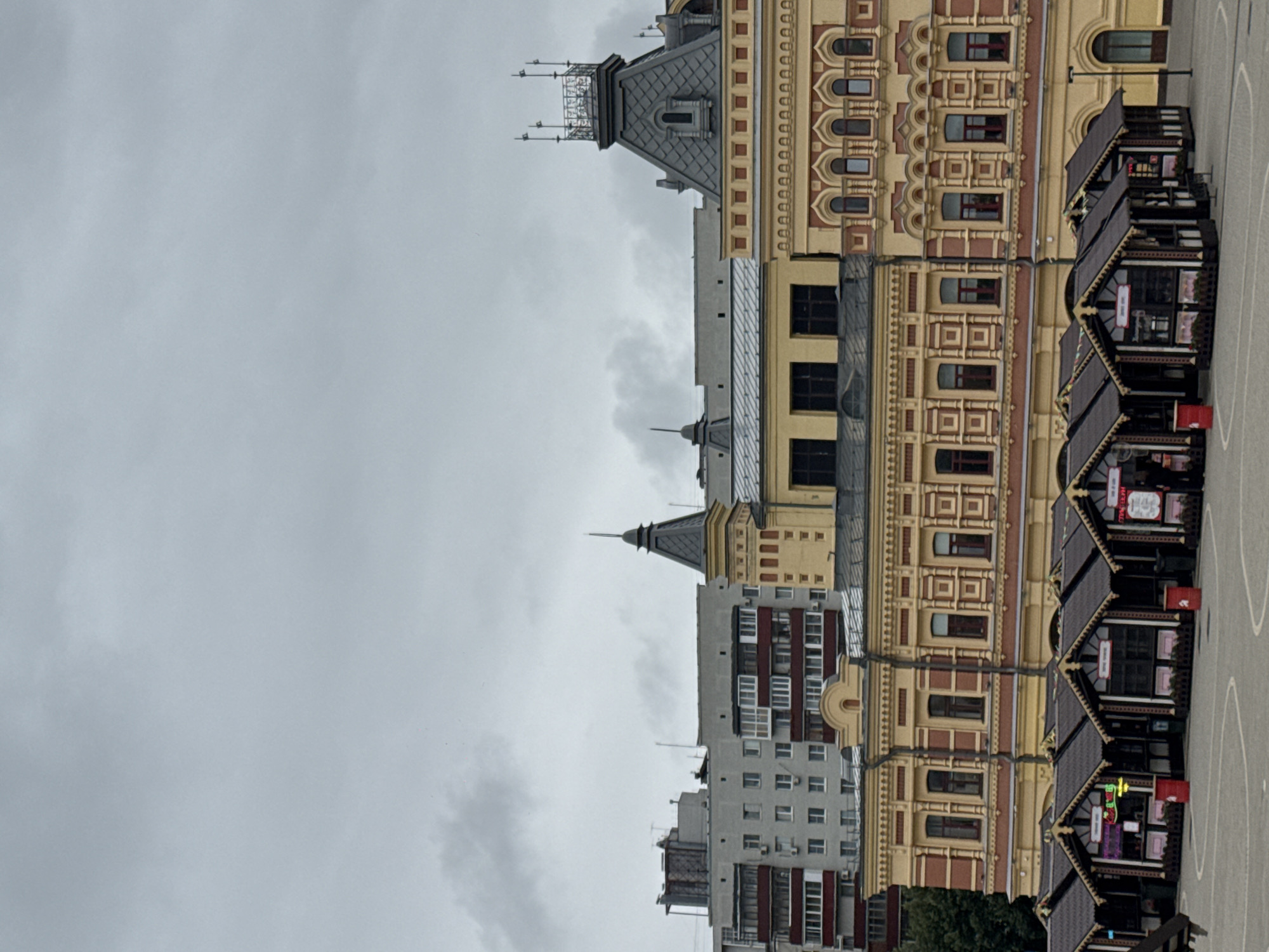 The image depicts a street scene in Nizhny Novgorod, Russia, on an overcast day. In the foreground, a row of small, dark-roofed market stalls with wooden structures are set up in a paved square. Each stall appears to be open for business, with some displaying signage, including neon lights in one stall. Red cylindrical trash bins are visible next to some of the stalls. In the mid-ground, a large, ornate building with yellow and red architectural details stands prominently. It features multiple windows and decorative elements. Behind this building, taller, more modern apartment buildings are visible. The sky is filled with heavy, gray clouds.