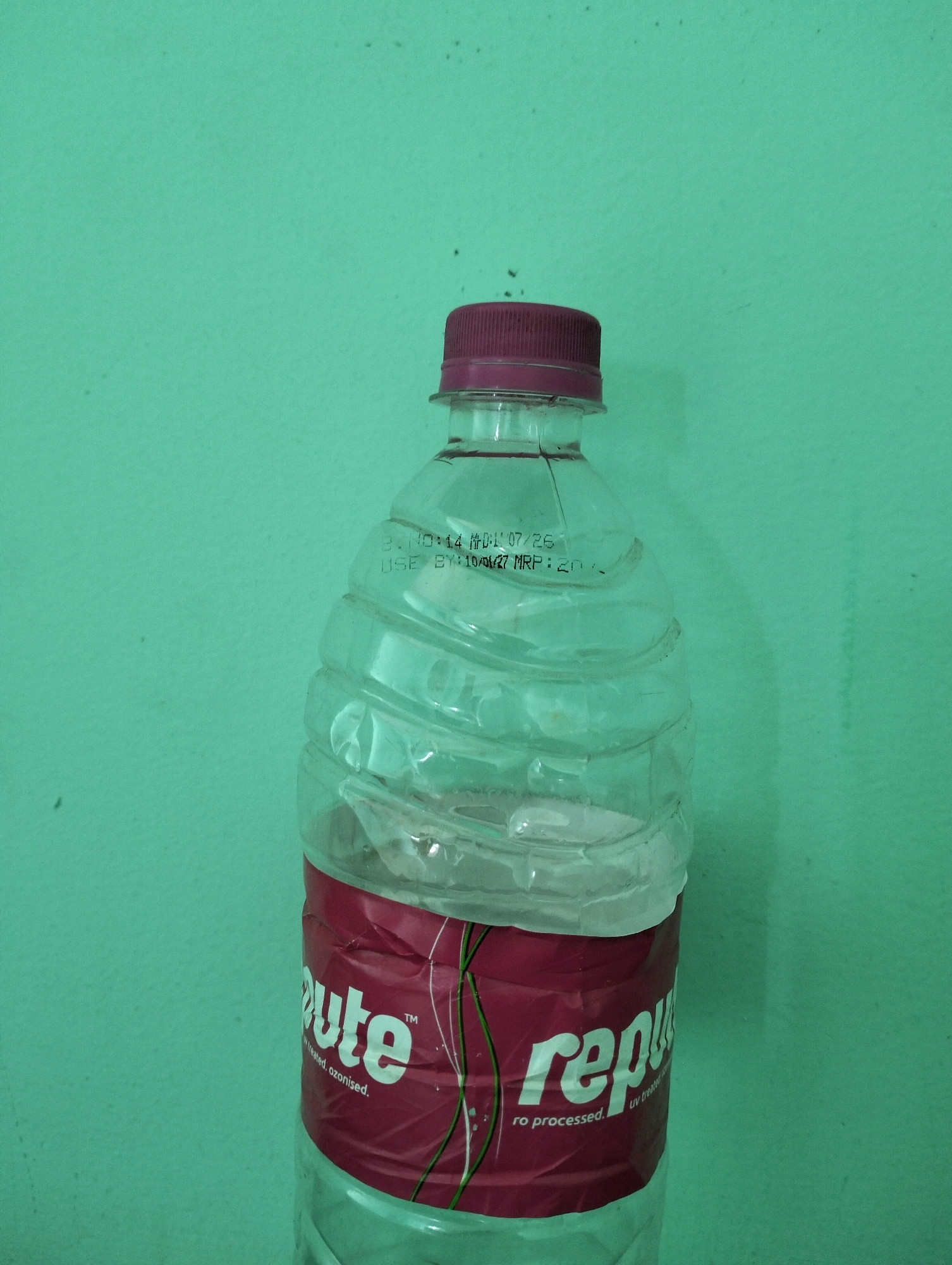 The image shows a clear plastic water bottle with a maroon label that says "re" on the left and "repu" on the right, suggesting the brand name is "Repute". The bottle has a maroon cap. There is a printed date and price on the bottle: "USE BY:10/07/26 MRP: 20/-". The bottle is placed against a plain, textured, teal-colored wall. The lighting suggests an indoor setting, likely a room with a single light source creating shadows. The image does not contain any information or visual cues that would allow for its location to be identified within Sriperumbudur, India, or any other city.