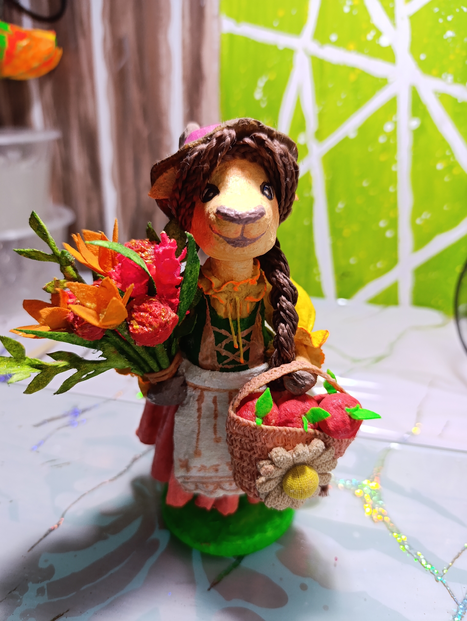 This is a close-up, eye-level, medium shot of a handmade figurine, likely crafted from clay or a similar material. The figurine depicts a character resembling a llama or alpaca, dressed in a rustic, historical costume. It wears a brown hat, a green bodice with orange lacing, a white apron with brown trim, and a pink skirt. The character has a braided brown hairstyle and a friendly, smiling expression.
The figurine is holding a bouquet of brightly colored flowers in its left hand, featuring orange and red blossoms with green leaves. In its right hand, it carries a small, woven basket filled with red apples and green leaves. The basket is adorned with a decorative flower made of beige yarn with a yellow center.
The background is softly blurred, creating a shallow depth of field that emphasizes the figurine. To the left, there are vertical, textured elements in muted brown and white, possibly representing curtains or a textured wall. To the right, there is a bright green background with a white geometric pattern, reminiscent of a leaf or a window pane, with subtle water droplets.
The figurine stands on a reflective surface, possibly a table, with a light blue and white marbled pattern. Scattered across the surface are small, twinkling lights, creating a festive or magical atmosphere. The lighting appears to be artificial, casting soft shadows and highlighting the textures of the figurine and its accessories. No text is visible in the image. The overall mood is cheerful and whimsical.