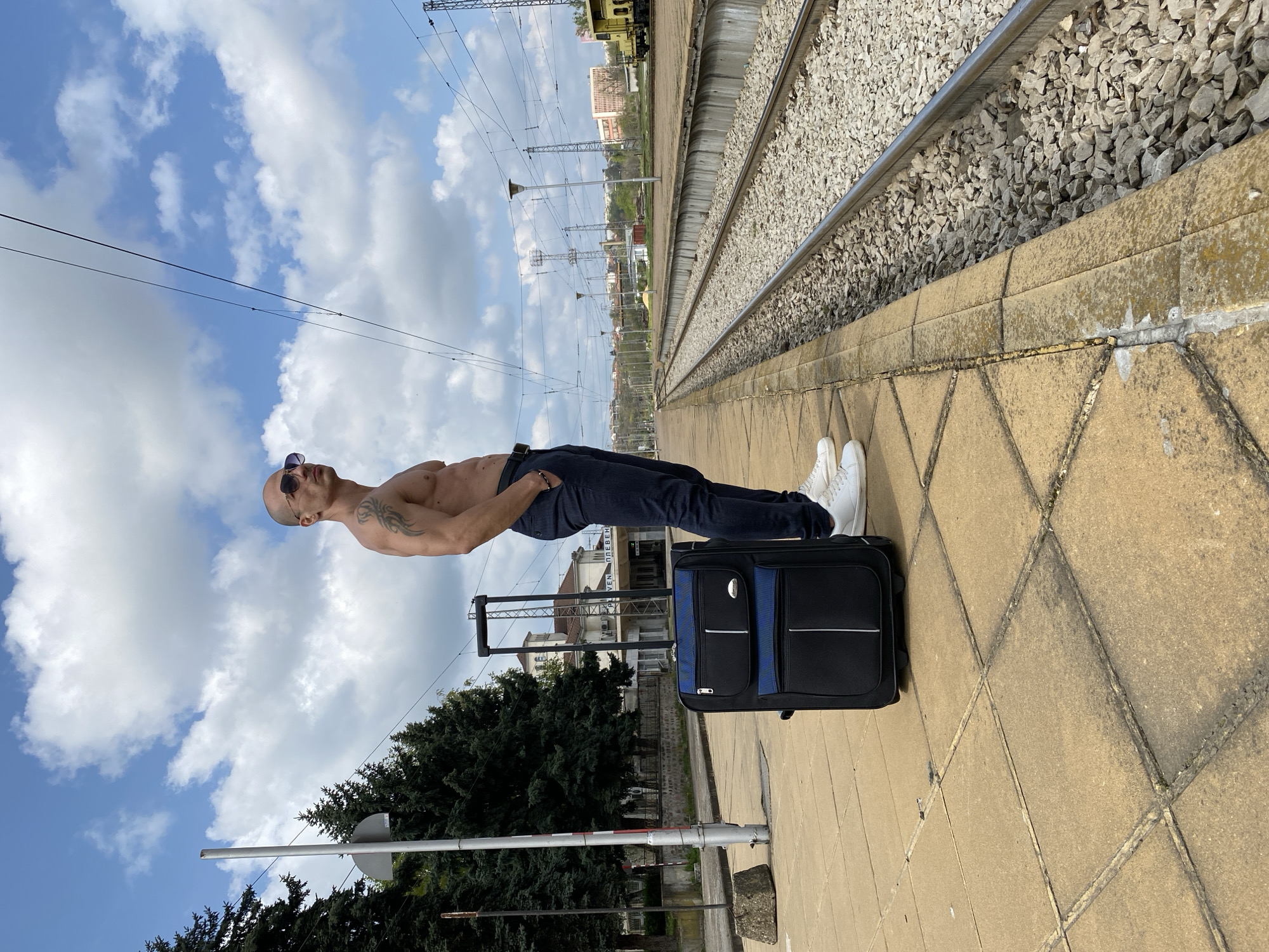 A man with a bald head and sunglasses, shirtless and wearing dark jeans and white sneakers, is lying horizontally with his feet supported by a black and blue suitcase with a handle, on a train platform in Pleven, Bulgaria. The platform is tiled with light brown stones. Railroad tracks run parallel to the platform. Buildings and greenery are visible in the background. The sky above features blue patches and clouds.