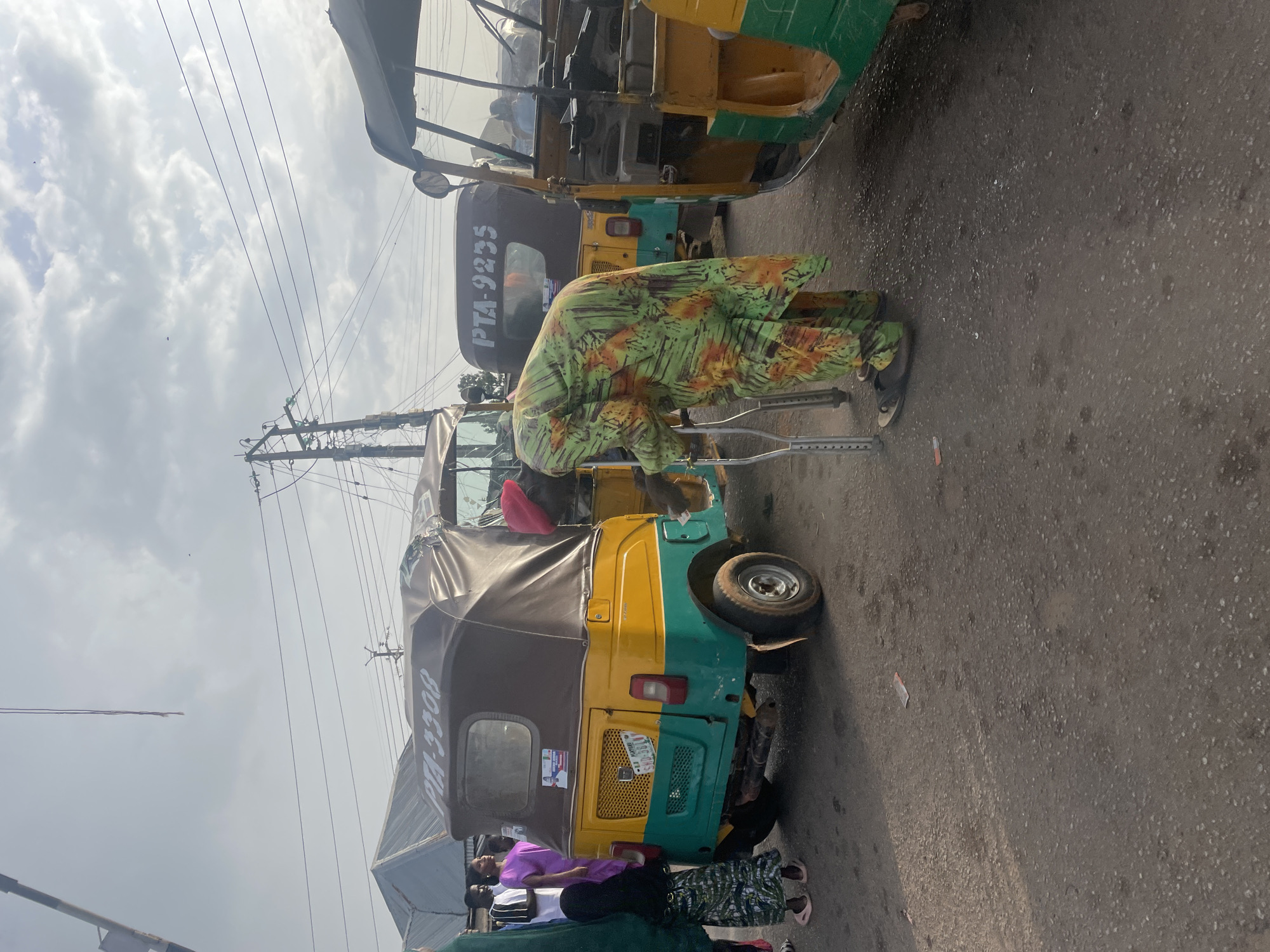 In Gura Top, Nigeria, an individual using crutches stands between two parked auto-rickshaws on an asphalt road. The person is wearing a patterned green and orange dress. The auto-rickshaws are painted in yellow and green. Several people are present near the vehicle in the foreground. Overhead are power lines against a cloudy sky.