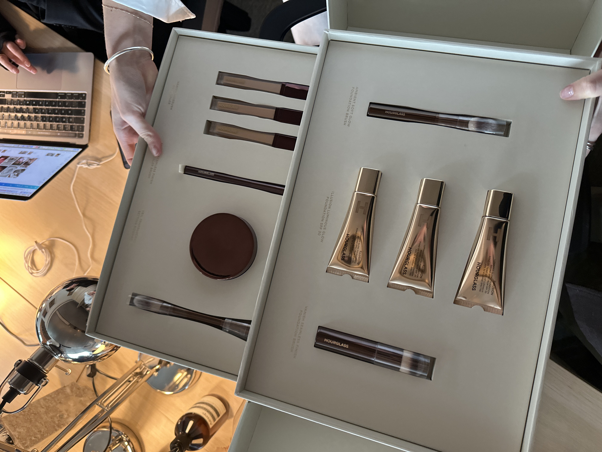 The image shows two open Hourglass makeup sets held by a person. The left set contains three brushes, a compact powder, and a blending brush. The right set contains a blending brush, three tubes of Hourglass foundation, and another brush. In the background, there's a laptop, a chrome desk lamp, and a bottle of Aesop hand balm on a wooden surface. While the image provides no explicit information about the location, it's possible that the photo was taken indoors at a house or in a professional setting like a beauty studio. The location cannot be confirmed without further information.