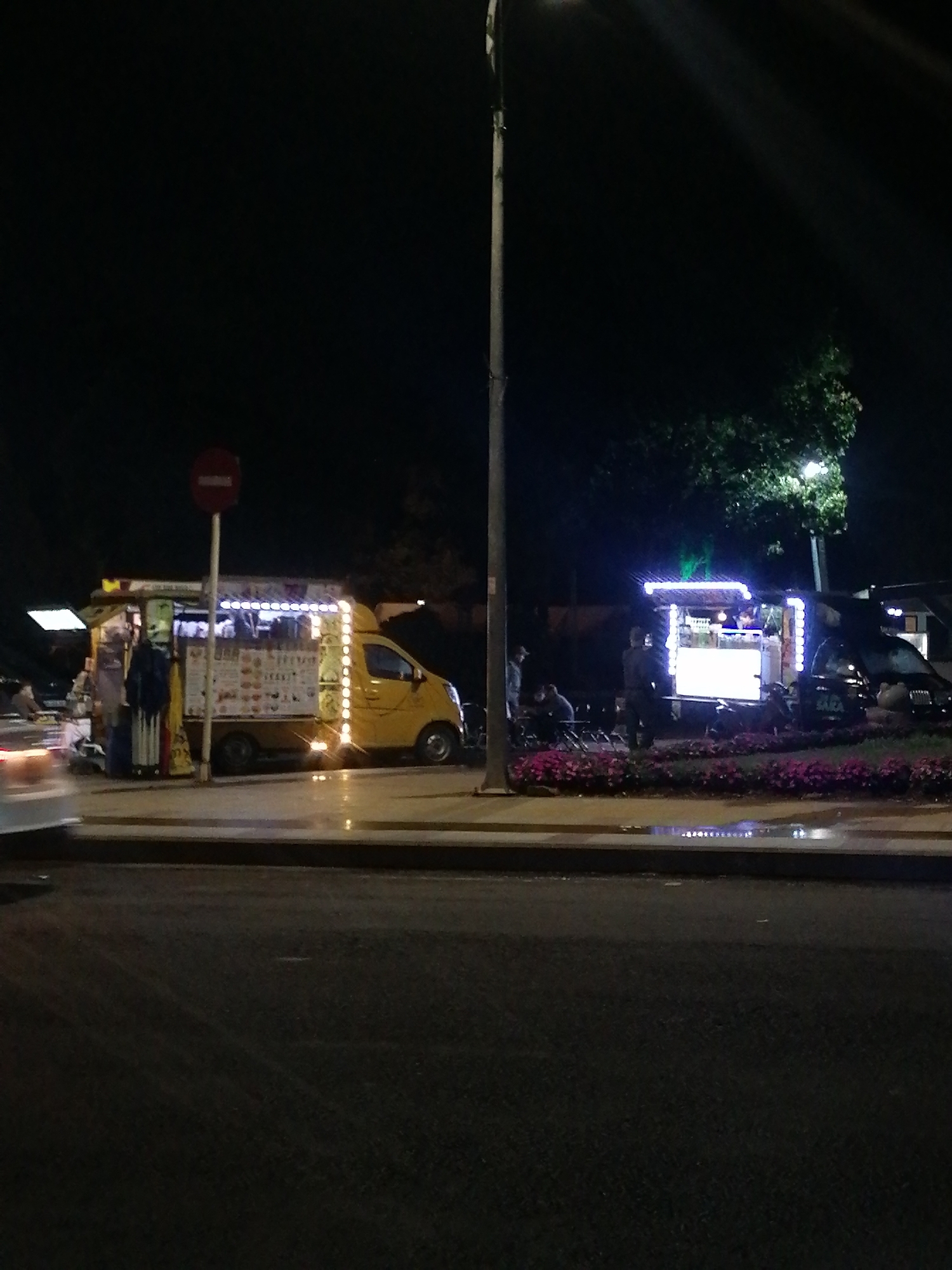 The image, taken at night, shows two food trucks located on a city street. The first truck is yellow with bright decorative lights and food menus displayed on the side. A red "no entry" traffic sign is nearby. The second food truck is dark in color, lit with blue and white lights, with tables and chairs near it. People are standing and sitting around both trucks. A flower bed with pink flowers separates the food trucks from the street. The street appears to be asphalt. A lamppost stands between the two trucks. Based on the known association of food trucks in Da Lat, Vietnam, it can be inferred that this scene may be there.