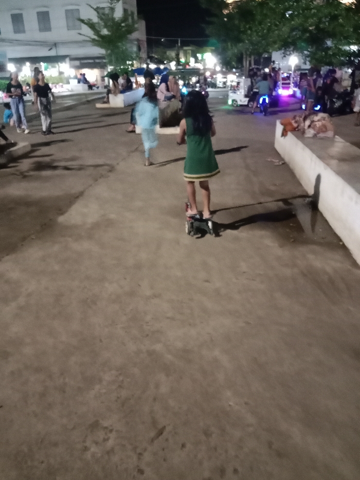 The image shows a young girl riding a scooter on a paved area in front of a building in Kaliwungu, Indonesia, located at -6.962 latitude and 110.253 longitude. The girl is wearing a green dress and her hair is pulled back from her face. The scooter is red and black and has a small, round, black seat. The girl is facing away from the camera and is riding towards the left. The pavement is grey and appears to be slightly wet. There are other people in the background, some of whom are walking and some of whom are sitting on benches. The building in the background is a two-story building with a white facade. There are trees on either side of the paved area and the background is obscured by darkness.