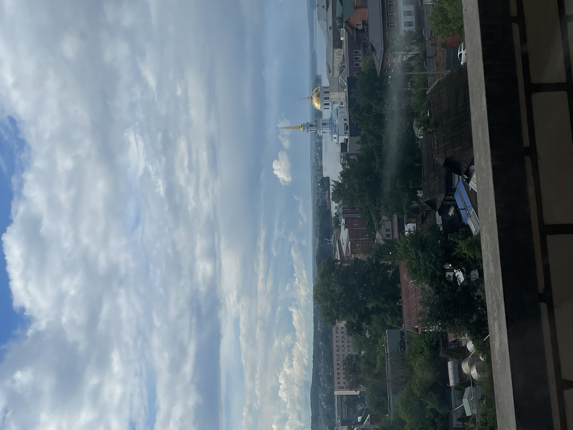 The image presents a panoramic daytime view of a cityscape, likely Votkinsk, Russia, captured from an elevated perspective, possibly a balcony or high window, indicated by a dark railing on the right.
The upper portion of the scene is dominated by a bright sky filled with various white and light grey clouds, some stretched and wispy, others more cumulus-like, suggesting fair weather.
The mid-ground reveals a townscape with numerous buildings, interspersed with lush green trees. A prominent architectural feature is a white building with a large golden dome and a slender golden spire, characteristic of a Russian Orthodox church, centrally positioned in the view. Beyond the urban structures, a wide body of water, possibly the Votkinsk Reservoir, stretches towards the horizon, where distant land or hills are faintly visible under the hazy sky. A white car can be seen on a street among the buildings.
The lighting indicates it is daytime, possibly late morning or early afternoon, with clear visibility. No specific human activity or visible text is discernible in the image.