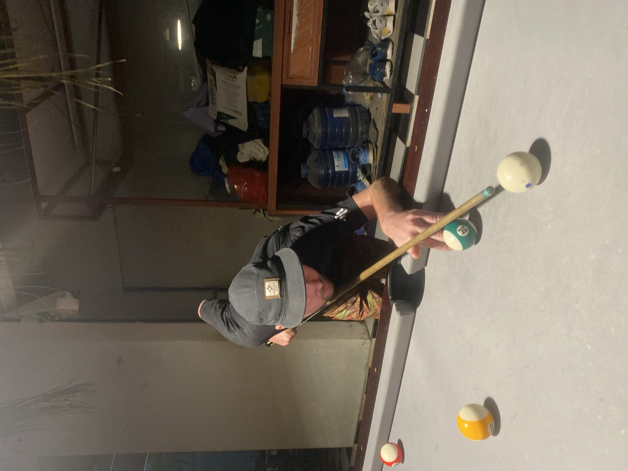 A person wearing a black long-sleeved shirt and a gray baseball cap is playing pool. The person is aiming a pool cue at a 14-ball, with the cue ball nearby. There are also a 9-ball and a red-and-white striped ball on the table. A shelf unit stands nearby and holds water bottles, a box, clothes, and shoes. The location is Mũi Né, Vietnam.
