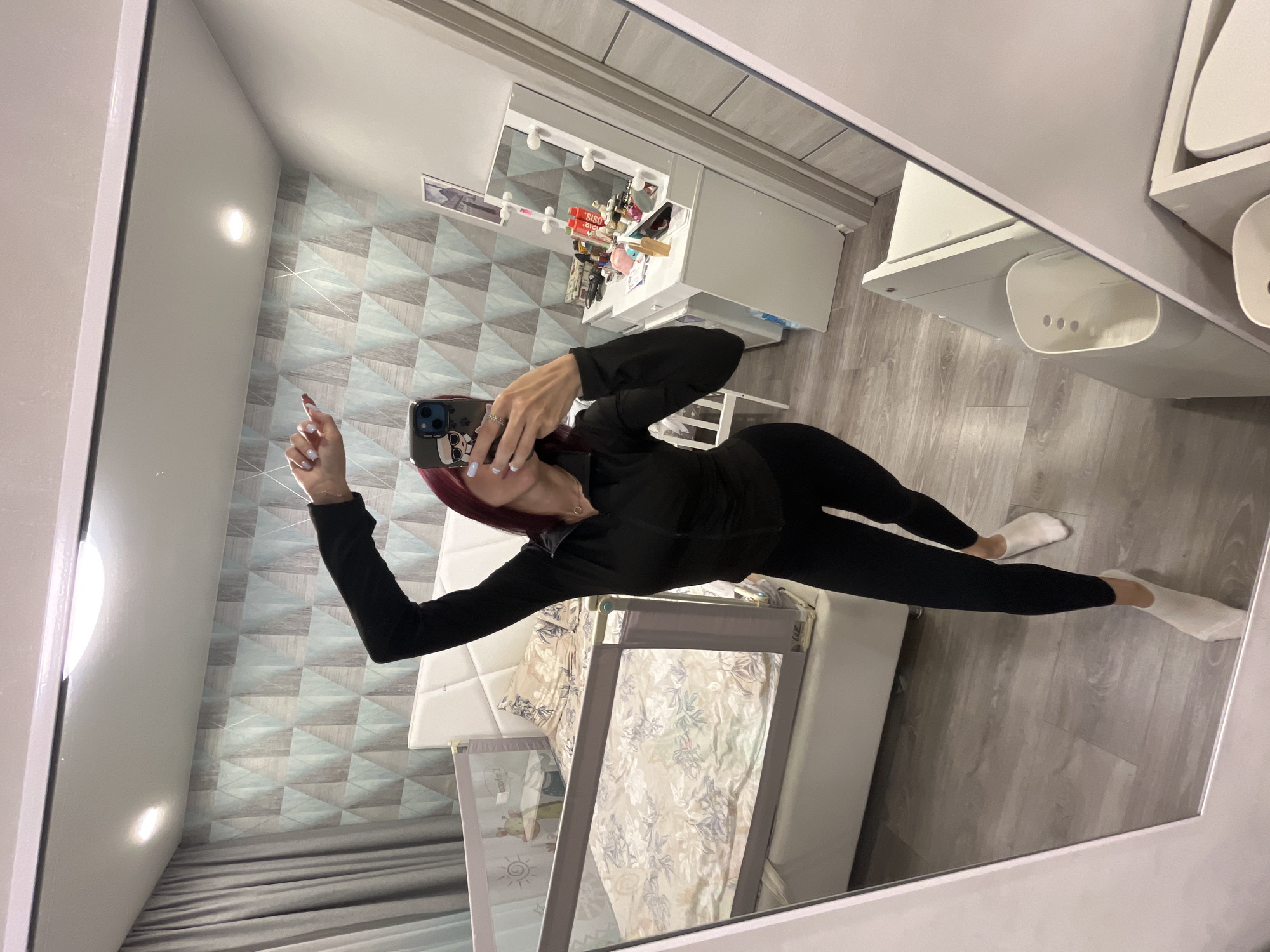 A woman with red hair takes a mirror selfie in what appears to be a bedroom. She is wearing a black zip-up jacket and black leggings, with white socks. Her left arm is raised, and she is holding her phone in her right hand. The room has patterned wallpaper, a large bed with a headboard, and a white vanity with a lighted mirror. A white baby crib is visible next to the bed. The flooring is light-colored wood laminate. This scene is not specific to Nizhny Novgorod, Russia.