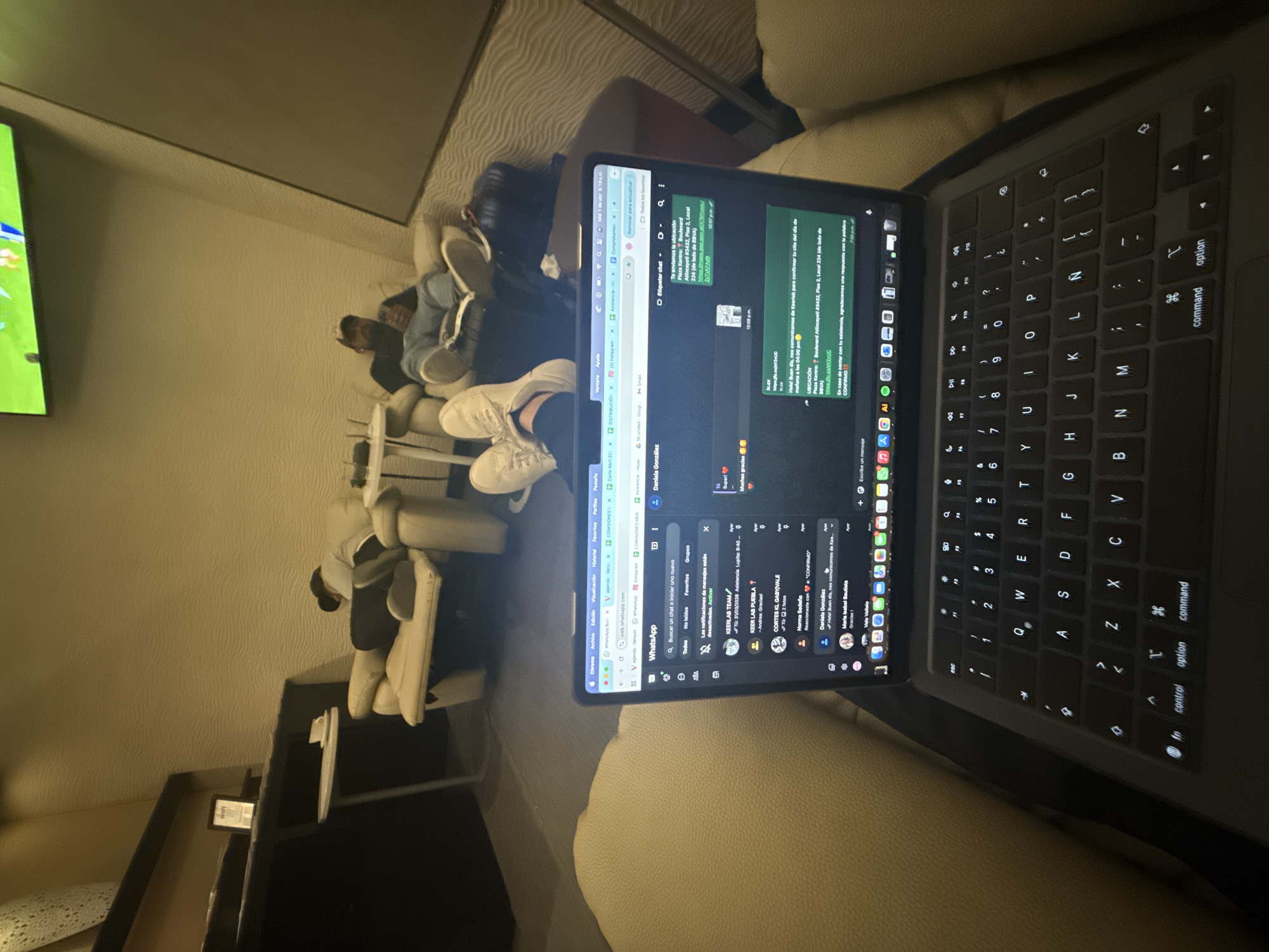 Here is a description of the image:
The image appears to be taken indoors, with a focus on a person seated on a cream-colored sofa. The individual is using a black MacBook laptop, with WhatsApp Web open on the screen. The screen displays a chat with location information for "Plaza Kentro: Boulevard Atlixcayotl #3432, Piso 2, Local 224." This location is likely in Mexico City, Mexico, as "Atlixcayotl" is a major boulevard in that city. The chat also indicates confirmation of an appointment for 4:00 PM.
In the background, other people are seated on similar cream-colored sofas, some facing a television screen. The environment suggests a lounge or waiting area, possibly in a hotel or office setting. A side table is visible with an item placed on it. The lighting appears dim, creating a relaxed atmosphere.