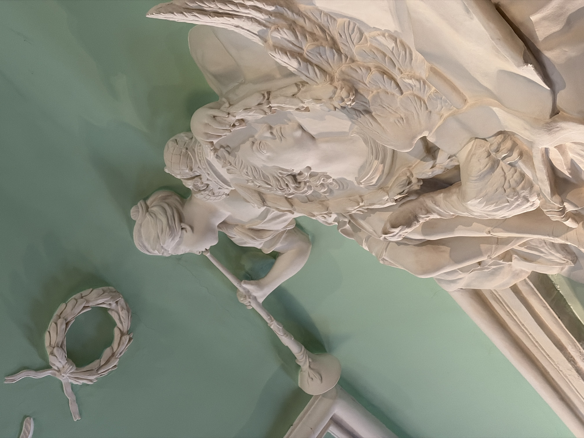 The image is a close-up of a stucco decoration on a ceiling in Saint Petersburg, Russia. The ceiling is light green. The stucco is white and features a woman with wings holding a horn and the face of a man with wings, with a laurel wreath off to the side. The figures are sculpted in a classical style. A partial white column is visible in the bottom right.