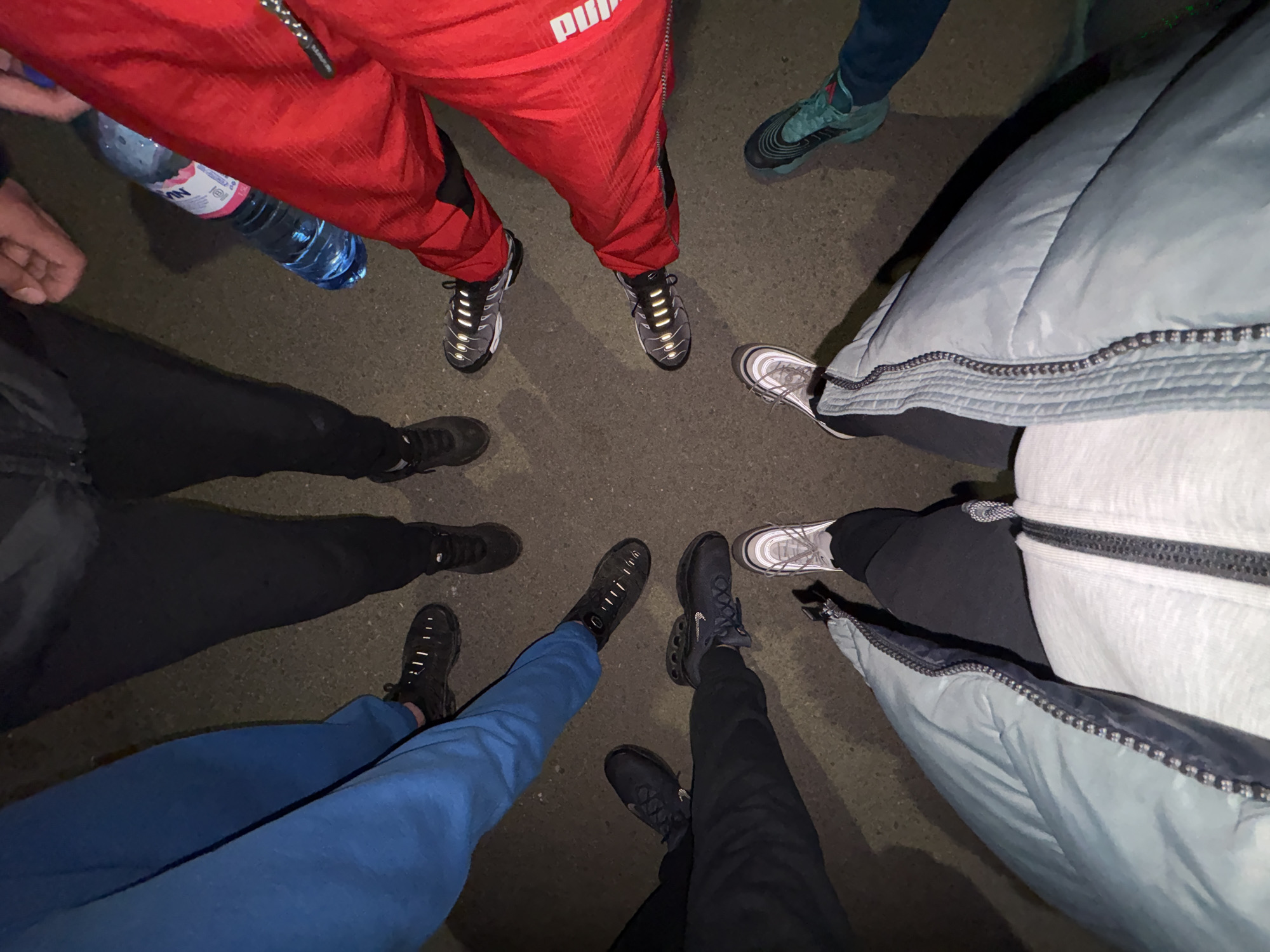 This overhead shot captures the lower halves of approximately six to seven individuals standing in a rough circle on a dark asphalt or concrete surface. The scene appears to be illuminated by artificial light, causing pronounced shadows and highlighting reflective elements on some of the footwear and clothing, suggesting it is nighttime or a low-light environment.
The subjects are dressed in casual athletic wear. One person in red track pants, clearly branded with "PUMA" on the leg, holds a plastic water bottle with a blue cap and a white label, on which "VIN" is partially visible. Other individuals wear black or blue track pants. A person on the right is seen wearing a light blue or grey puffer jacket over a lighter garment.
A variety of athletic shoes are visible, predominantly Nike models. Several pairs of Nike Air Max TN sneakers, recognizable by their distinct design and some with reflective silver accents, are present. Other footwear includes a pair of Nike Air Max 97s with silver and white detailing, and several pairs of dark, chunky-soled sneakers.
The activity depicted is a gathering, with the individuals standing close together with their feet pointing towards a central point. There are no direct cues about emotions or specific activities beyond standing and perhaps conversing. The location is an unidentifiable outdoor or paved area.