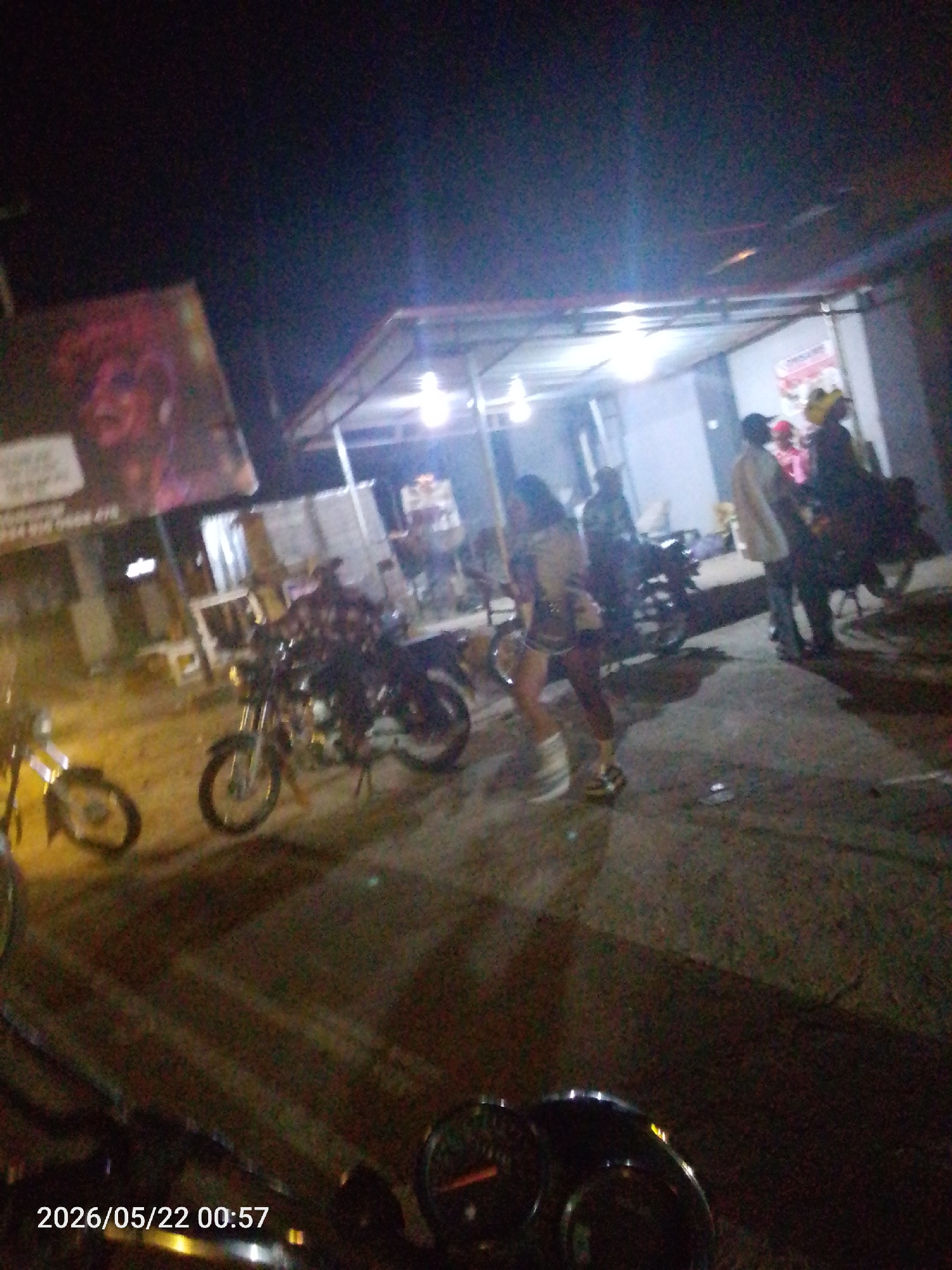 The image shows an outdoor scene at night in Lokoja, Nigeria. Several people are standing near a building with a corrugated metal roof and open sides, which appears to be a roadside business or gathering place. Several motorcycles are parked nearby. A person in shorts and a jacket is walking. A large billboard with a face is visible to the left. The image has a timestamp indicating it was taken on 2026/05/22 at 00:57. The location appears to be a gathering spot with transportation.