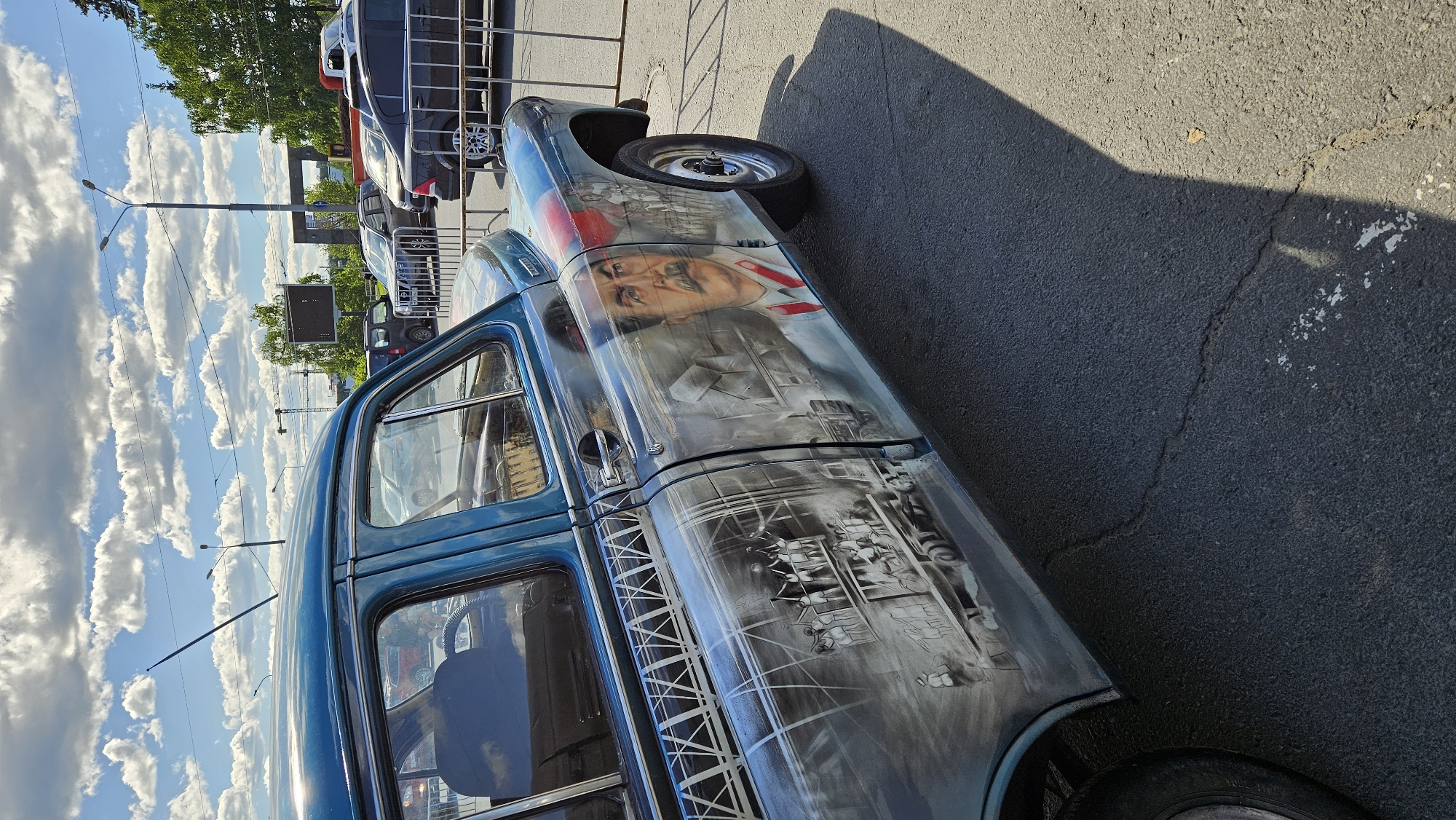 A teal-colored vintage car is parked outdoors, its side adorned with an airbrushed mural. The mural depicts a portrait of Joseph Stalin on the rear door, with a scene of workers and a train on the front door and fender. The car is positioned on an asphalt surface with visible cracks. In the background, other vehicles are visible, along with trees, streetlights, and a cloudy sky. A metal fence runs alongside the car. The image appears to be taken during the daytime.