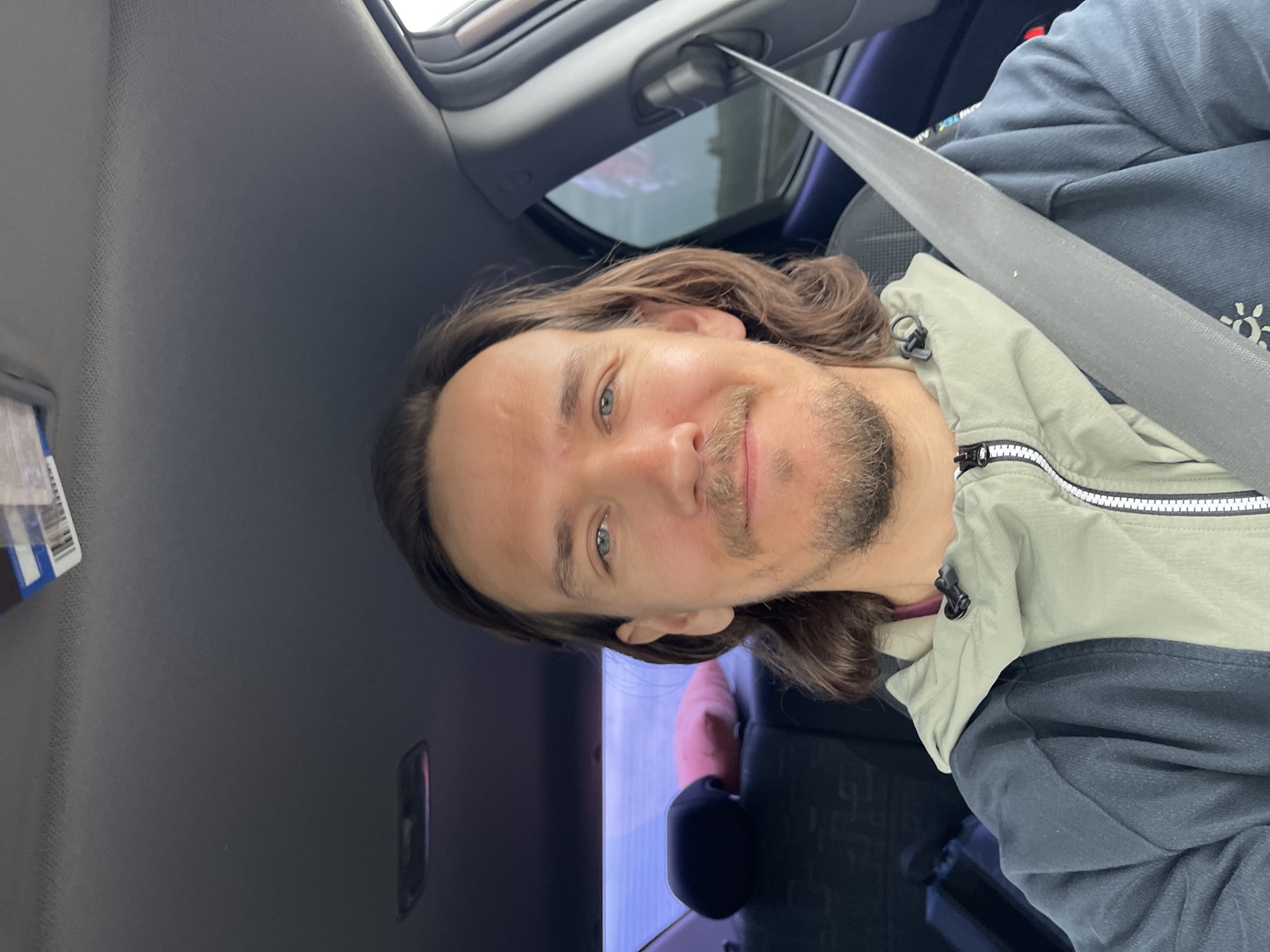 A middle-aged man with shoulder-length brown hair and a beard is in the front seat of a car, wearing a light green and dark blue jacket and a seatbelt. He is looking directly at the camera with a slight smile. The car has a sunroof. The exact location in the city of Niva, Russia is not identifiable from the provided image.