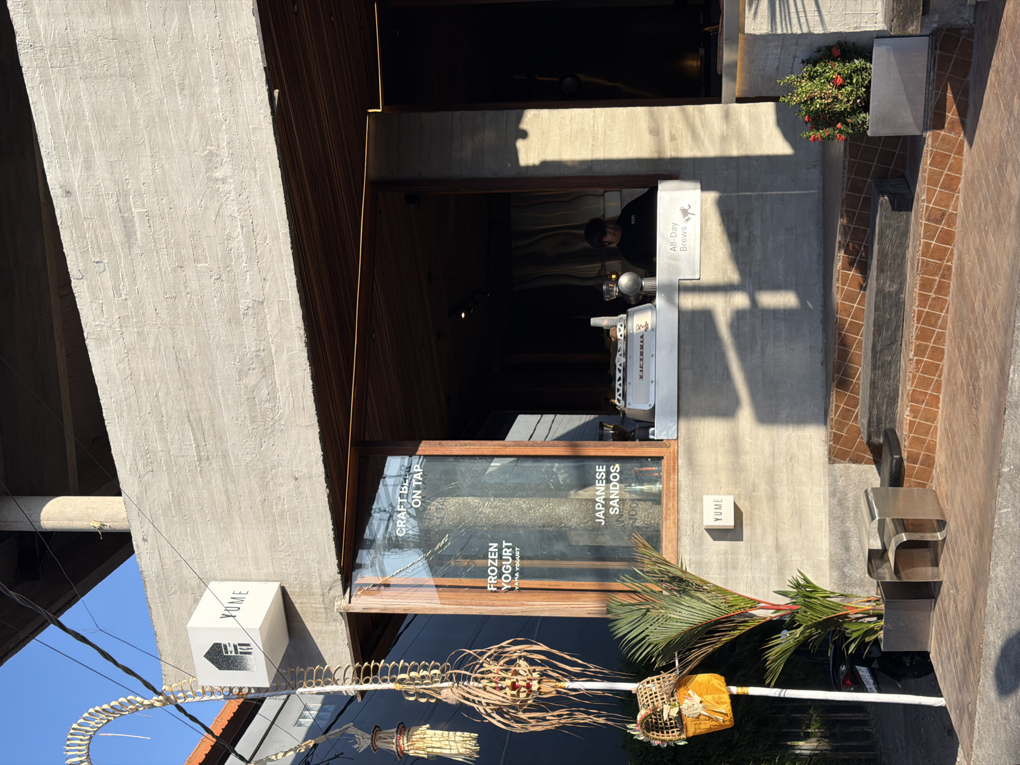 This image shows the exterior of a cafe named "YUME" located in North Kuta, Indonesia. The building features a minimalist concrete facade with wooden accents. A cubic sign with the "YUME" logo is mounted on the upper concrete section. A large glass window reveals the interior, where a barista is visible behind a counter. Text on the window indicates offerings such as "CRAFT BEER ON TAP," "FROZEN YOGURT," and "JAPANESE SANDOS." In front of the cafe, there are potted plants, a metallic bench, and a motorbike parked to the left. Traditional Balinese decorative elements, made of dried leaves and straw, hang from a pole on the left side of the entrance. The scene is bathed in the warm light of what appears to be late afternoon.