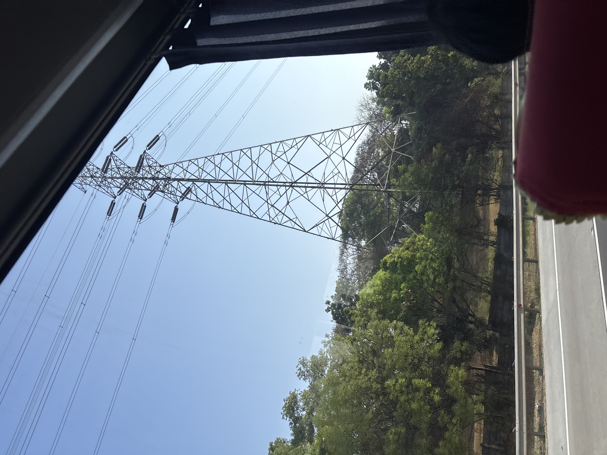 A tall, lattice-style transmission tower stands against a clear blue sky, supporting multiple power lines that stretch across the frame. Dense green trees and foliage surround the base of the tower, with a concrete barrier and guardrail separating the area from a paved road in the foreground. The view appears to be from a moving vehicle, possibly a bus, with the edge of a dark curtain and a hint of a red seat visible in the lower right corner. The scene is located in Karawang, Indonesia, a region known for its industrial areas and power infrastructure.