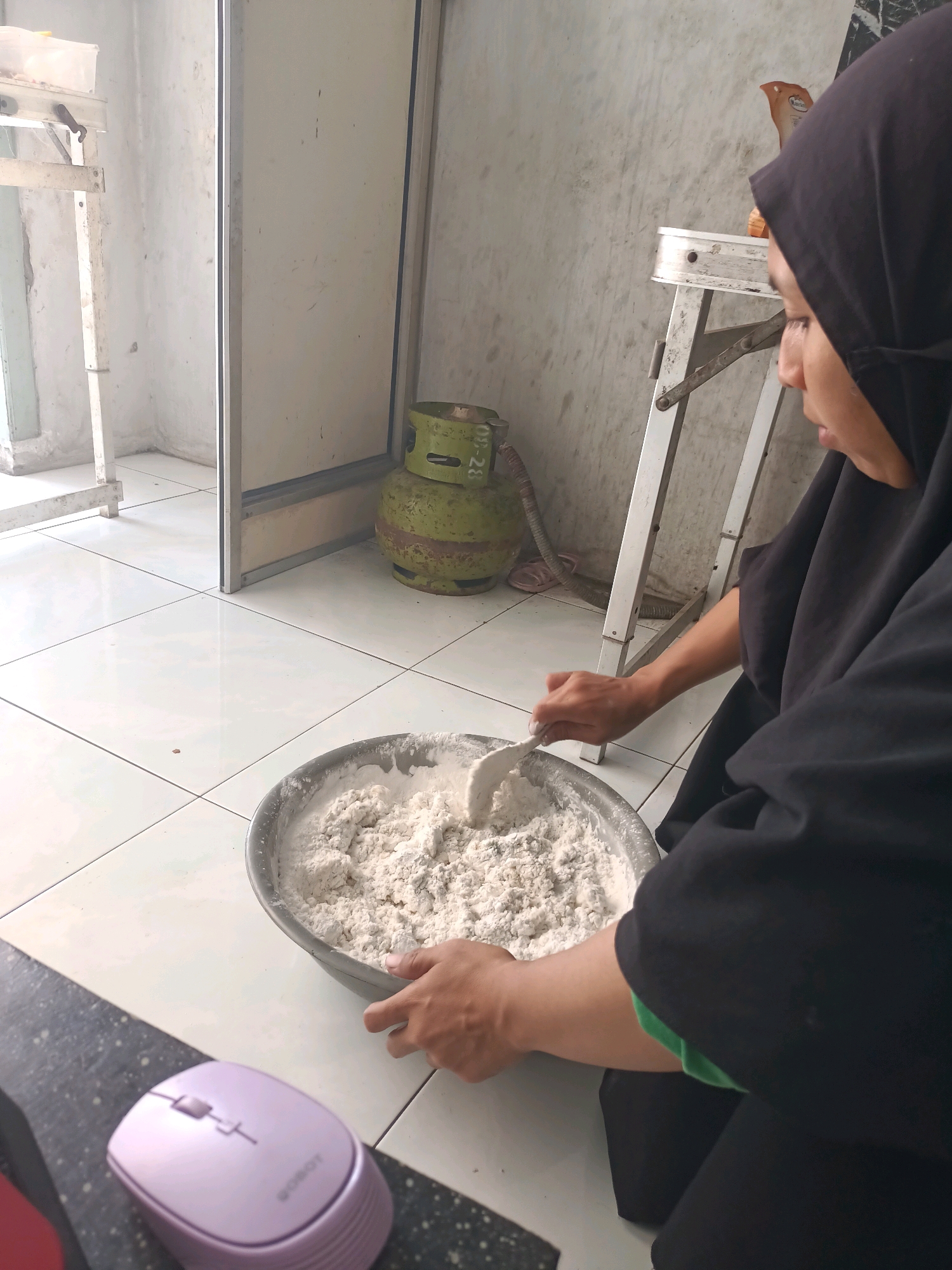 In a domestic setting in Badas, Indonesia, a person wearing a black hijab is preparing dough in a metal bowl. The individual is using a spatula to mix the dry ingredients, which appear to be flour and possibly other powdered substances. The bowl is placed on a tiled floor. In the background, a green gas cylinder is visible, along with a metal shelf unit. To the left, a doorway leads to another part of the room, with a glimpse of a white wall and what appears to be a metal frame. A light purple computer mouse is in the foreground, to the left of the bowl. The scene is lit by natural light, casting soft shadows.