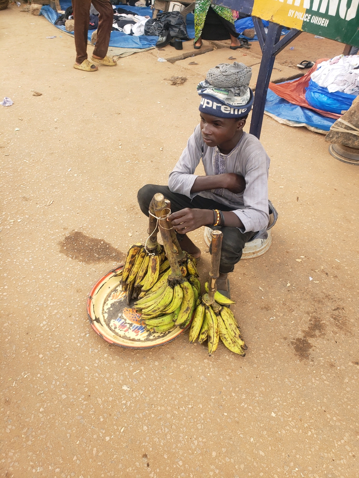A young male, dressed in a light grey long-sleeved tunic with embroidered trim and black pants, sits on a white bucket by the side of a road in Sabon Tasha, Nigeria. He wears a grey and white patterned cap atop his head. He is looking off to his right. In front of him is a platter of green and yellow bananas. He appears to be selling the bananas. In the background, there are blue and orange tarps spread out on the ground, with piles of clothing and bags. A sign with "BY POLICE ORDER" is visible. The ground is dusty and uneven, with scattered debris.