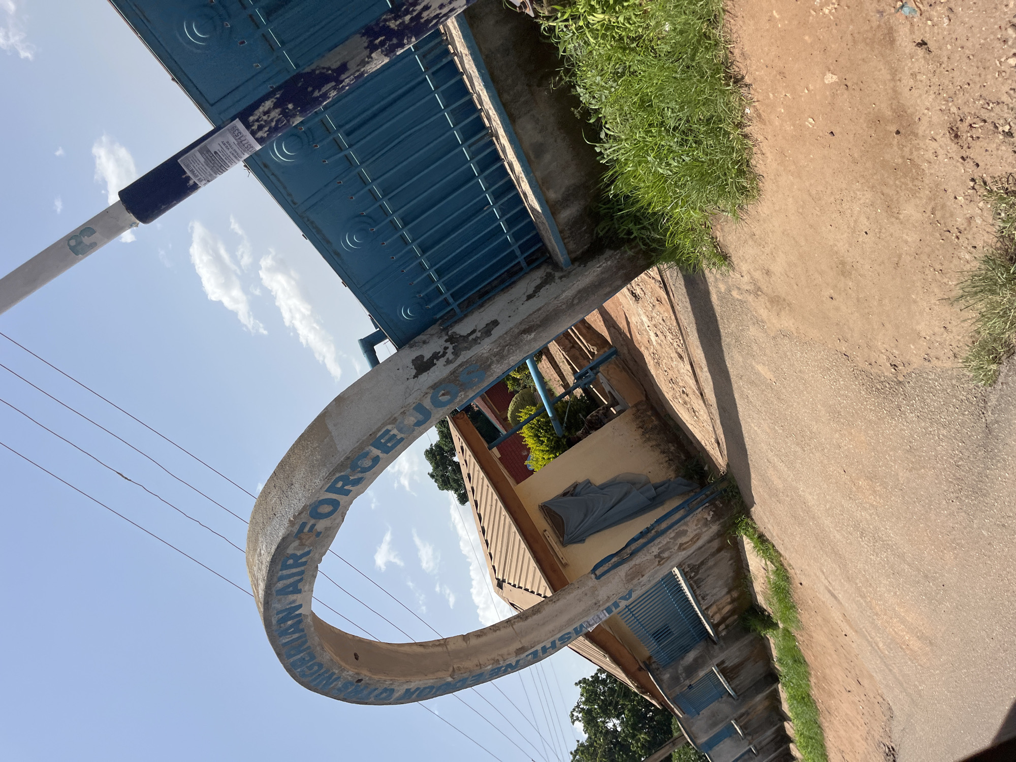 The image shows an outdoor scene at an entrance. A concrete archway with blue lettering that reads "NIGERIAN AIR FORCE" is visible. Beyond the arch, a blue metal gate stands closed. To the left of the gate, a light-colored building with a brown roof is partially visible. There is a dirt path leading to the gate and a grassy area to the right. The sky above is blue with scattered white clouds. The scene appears to be in Gura Top, Nigeria. There are no people or animals in the image. The overall impression is one of an official entrance to a military facility.