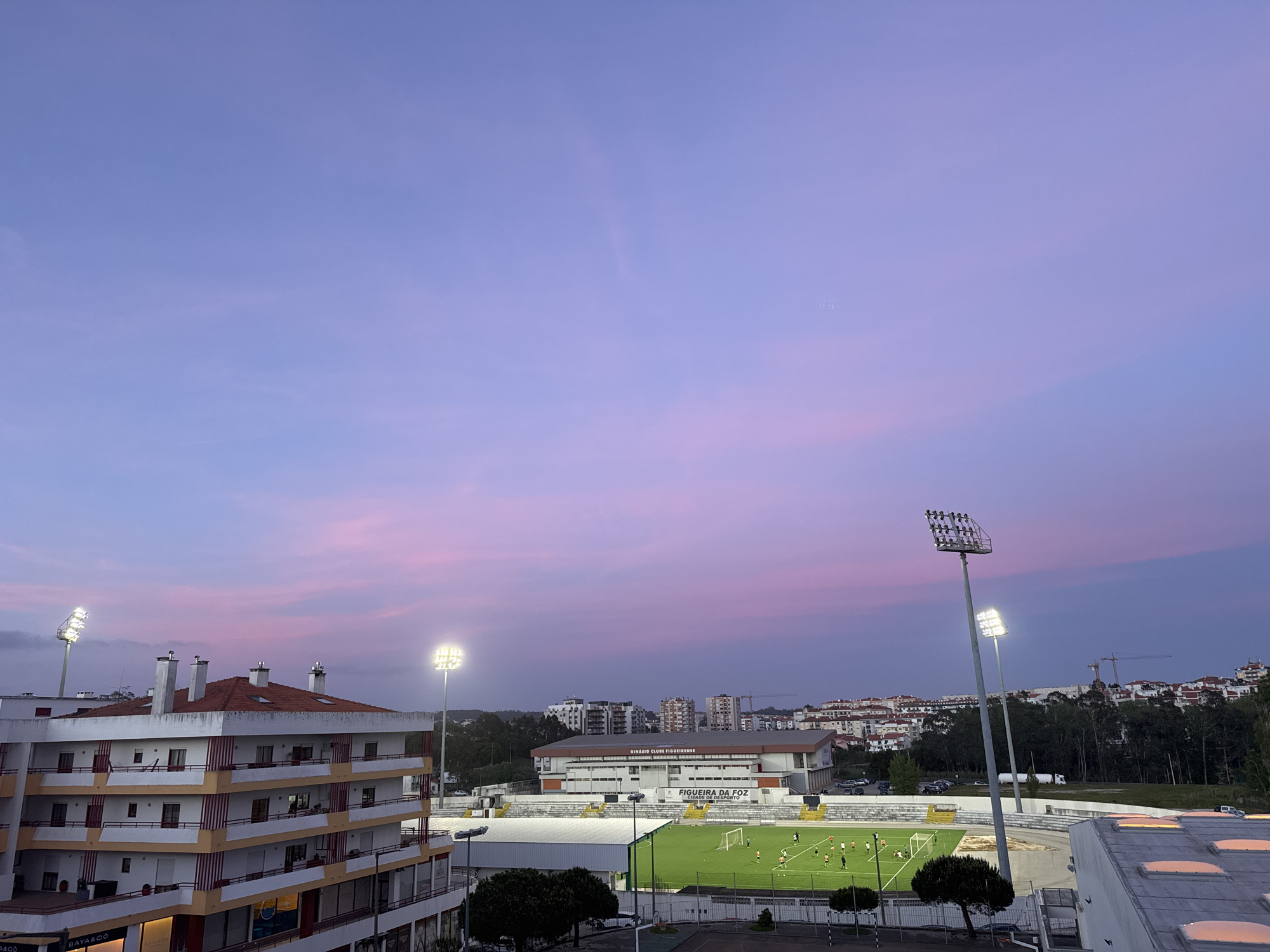 The image presents an elevated view of Figueira da Foz, Portugal, during twilight. The most striking feature is the sky, which transitions from light blue at the top to vibrant shades of purple and pink closer to the horizon, with some faint, wispy clouds adding texture.
In the mid-ground, a brightly lit football (soccer) pitch is the focal point, where a group of players are actively engaged in a game or training session. The green turf and white lines are clearly visible, as are the two goalposts. Surrounding the pitch are stadium stands and structures. Tall floodlight towers, with several lights illuminated, cast a strong artificial glow over the field, contrasting with the soft natural light of dusk.
Behind the stadium pitch, a large building bears the text "GINASIO CLUBE FIGUEIRENSE" above and "FIGUEIRA DA FOZ" below, clearly identifying the location and a local sports club. Further in the background, a residential or commercial cityscape extends, featuring numerous buildings of varying heights and styles, interspersed with areas of dense trees. Construction cranes are visible in the distance, indicating ongoing development.
In the foreground, to the left, a multi-story building with a red-tiled roof and balconies is partially visible. On its ground floor, the text "BAY & CO" is discernible. The scene captures a moment where evening sports activity unfolds under a beautifully colored sky, blending urban environment with natural aesthetics.