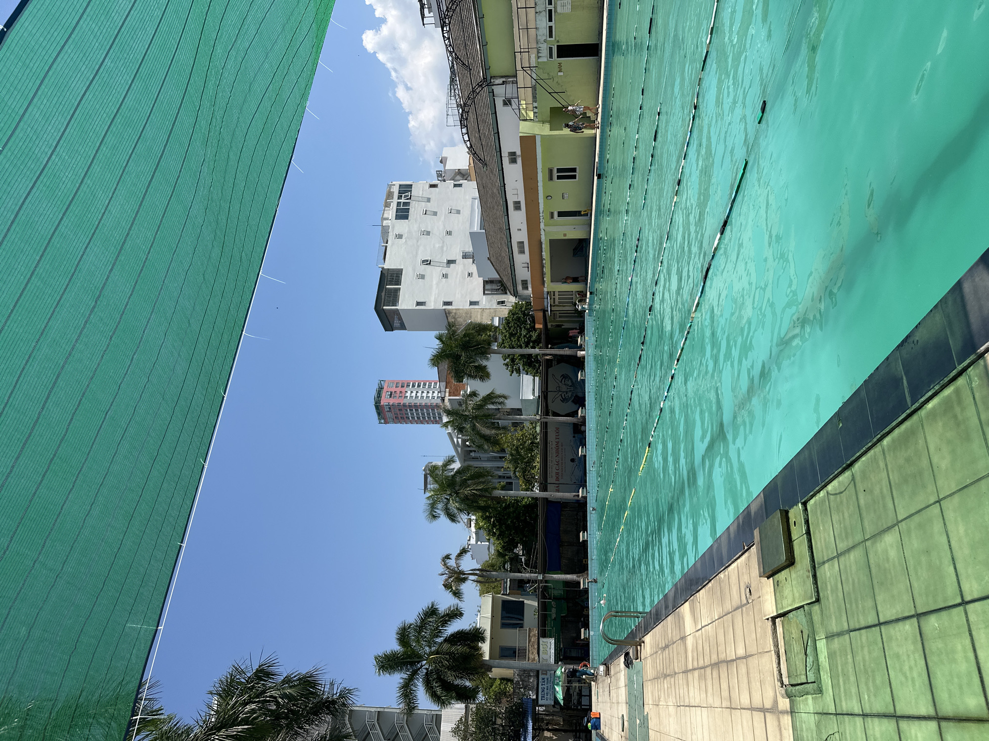 This is a photograph of a swimming pool facility in Nha Trang, Vietnam. The pool is outdoors, with multiple lanes marked by ropes and buoys. A green sunshade is stretched over the top of the image, partially obscuring the bright blue sky. Palm trees and several buildings, including a tall, pink-and-white skyscraper in the distance, surround the pool. The pool area appears to be a public facility, with tiled pathways and some swimmers in the water. There are also several people walking around the pool. The overall impression is of a sunny day at an urban swimming pool.