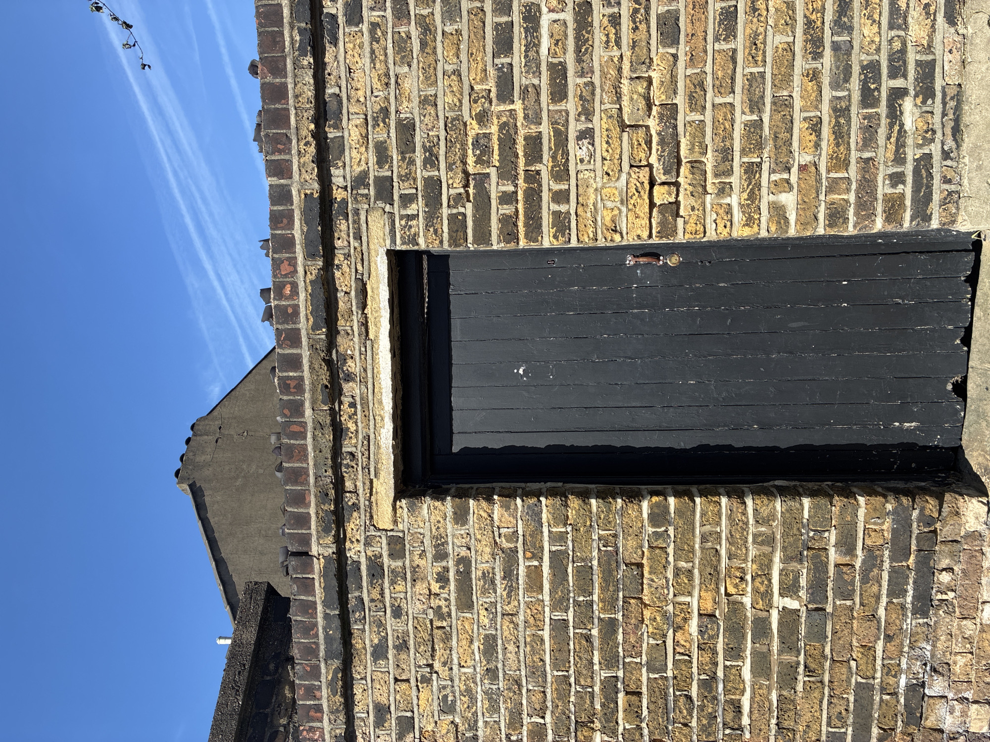 This image shows the exterior of a building, possibly a shed or small structure, constructed with yellow brick. A black door is set into the brick wall. The surrounding brickwork exhibits signs of weathering and discoloration. Above, a portion of a roof is visible against a clear blue sky with contrails. The image suggests a location in an older urban environment.
Given the style of the brickwork, this is likely to be located in the United Kingdom, and possibly London.