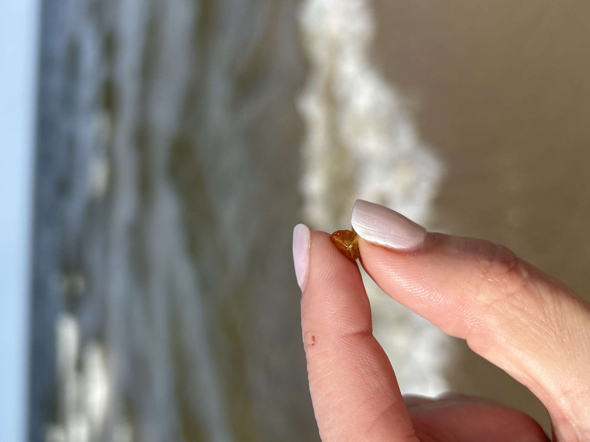 A person holds a small, irregularly shaped, amber-colored stone between their thumb and index finger. The fingernails are manicured with a light pink, iridescent polish. In the background, out of focus, is a blurred view of a sandy beach with ocean waves. Given the presence of amber, the scene is likely located somewhere along the Baltic Sea coast, which may be Gdańsk, Poland.