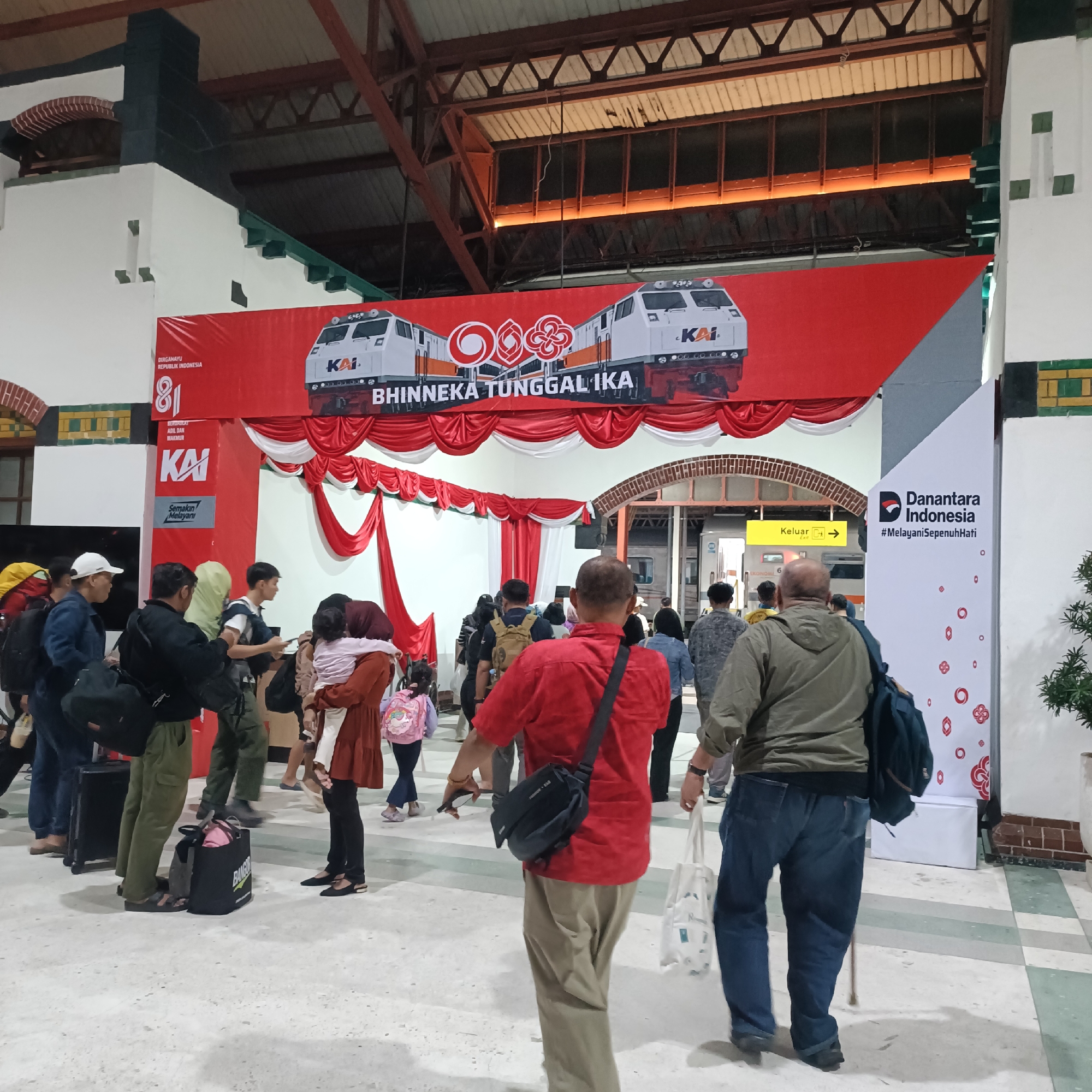 The image depicts a scene inside a train station in Semarang, Indonesia, likely the historic Semarang Tawang Station, given its distinctive architecture and the celebratory banner. A large red banner hangs prominently, displaying the KAI (Kereta Api Indonesia) logo and text related to "Dirgahayu Republik Indonesia" and "Bhinneka Tunggal Ika," suggesting a national celebration or anniversary event. Two train engine graphics are featured on the banner.
In the foreground and midground, a crowd of people is gathered, appearing to be travelers. Many are carrying luggage, including backpacks, duffel bags, and rolling suitcases. The people are of various ages and genders, dressed in casual attire. Some individuals are walking, while others appear to be waiting or queuing. A woman in a reddish-brown dress is holding a young child.
The environment is that of an indoor public space, characterized by a high ceiling with exposed beams and lighting fixtures. The floor is tiled, with a pattern of light and dark squares. Architectural elements, such as white pillars with green trim and decorative tiling, are visible. A yellow sign with the word "Keluar" (Exit) and an arrow indicates an exit point. To the right of the banner, a white panel displays the "Danantara Indonesia" logo and the hashtag "#MelayaniSepenuhHati" (Serving with All Heart). The overall atmosphere appears to be busy and communal, indicative of a transit hub during an event.