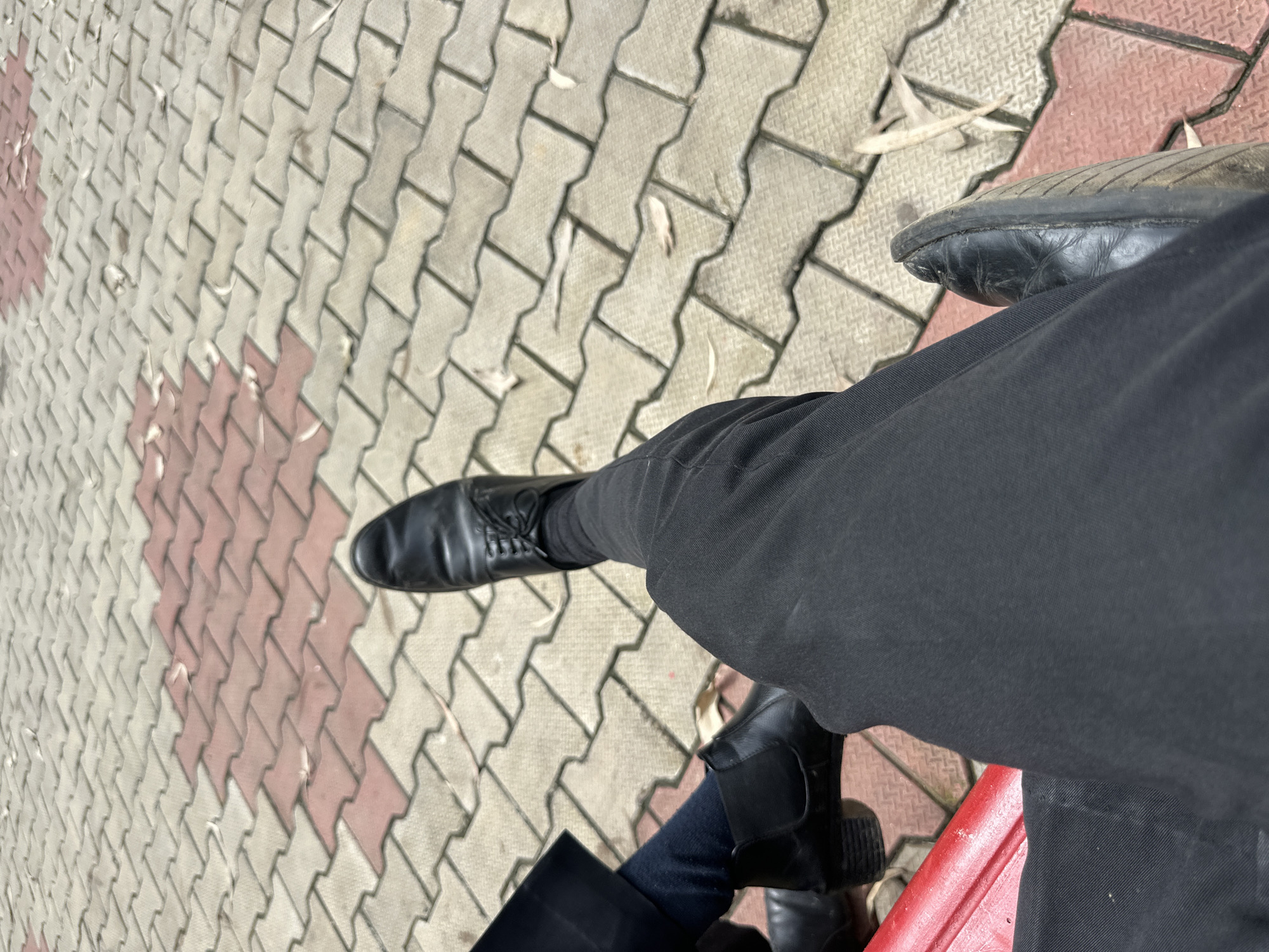 The image shows a close-up view of two pairs of legs and feet wearing black dress shoes and dark pants, resting on a patterned paved surface. The paving stones are interlocking and colored in alternating shades of beige and red, with fallen leaves scattered across them. The perspective is from above, looking down at the legs and feet. One person's legs are positioned in the foreground, with their black dress shoe prominently visible. Another person's legs are to the left, showing black boots and dark socks. A red bench is partially visible in the lower left corner. The scene appears to be an outdoor urban setting, possibly a park or public walkway, in Unknown, Unknown.