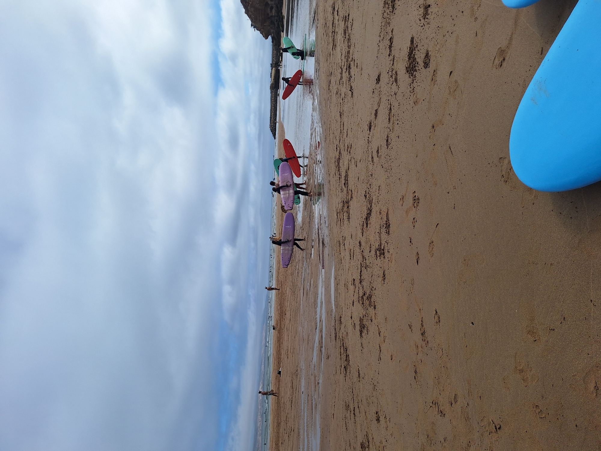 The image captures a wide, sandy beach in Imsouane, Morocco, under a cloudy sky. The foreground is dominated by the wet sand of the intertidal zone, with a few scattered footprints. Further back, a group of people are visible near the water's edge, some carrying surfboards. Specifically, to the left, a person in a dark swimsuit carries a light purple surfboard. To their right, another person in dark clothing holds a pink surfboard, followed by someone with a red surfboard. In the middle ground, two individuals are in shallow water, each holding a surfboard; one has a red board, and the other, a green one. Further out, more people and surfboards are visible in the ocean, along with the distinctive Imsouane bay and its natural rock formations, indicating the location is likely near the famous surf spots. The overall scene depicts a typical day at this Moroccan surf town, with surfers preparing to enter or exiting the water.