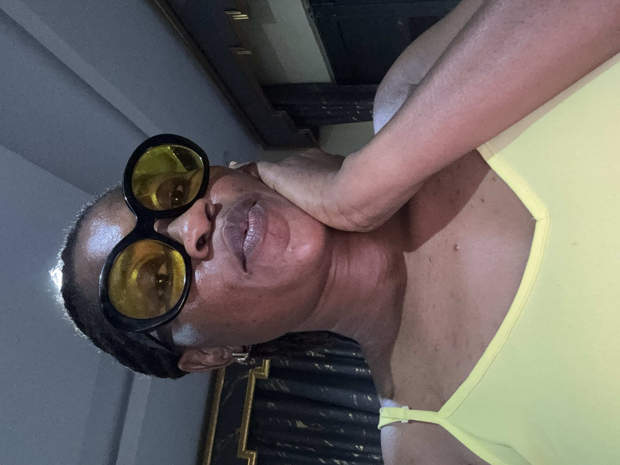 In a medium shot, a Black woman wearing large, round, yellow-tinted sunglasses and a yellow tank top is looking towards the camera. Her left hand is resting on her chin and cheek. Her hair is braided and pulled back. The background appears to be indoors with dark curtains visible behind her left shoulder, and a wall or doorway to her right. The lighting is dim, with a bright light source reflecting on her forehead. The scene is consistent with an indoor selfie taken in a residential setting in Ajara, Nigeria.