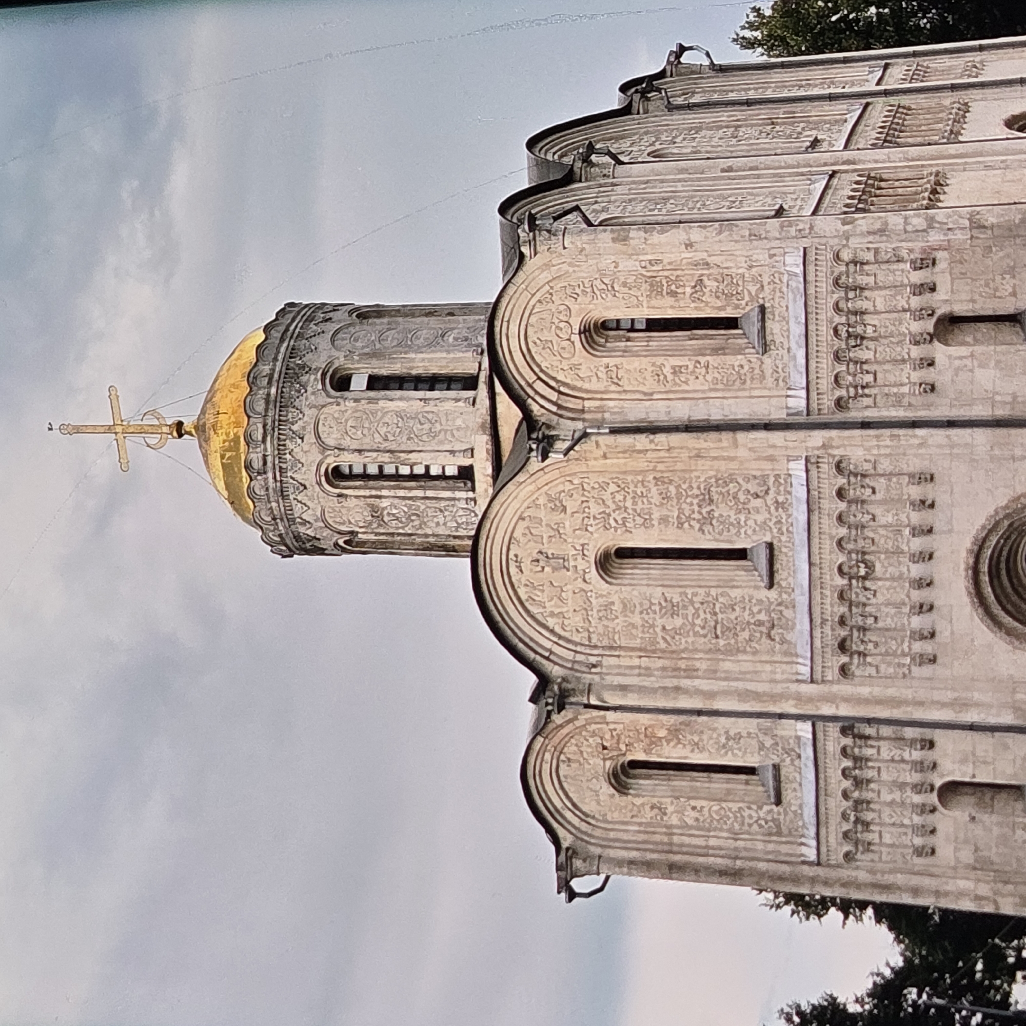 The image shows a close-up, low-angle view of the Dormition Cathedral in Vladimir, Russia. The cathedral, constructed of white limestone, features intricate carvings and a prominent golden dome topped with a cross. The sky above is overcast with gray clouds. The foreground is dominated by the facade of the cathedral, with its arched windows, decorative friezes, and a series of small arched niches. Dark green trees are visible on the left and right edges of the image, suggesting an outdoor setting. The architectural style is characteristic of ancient Russian churches.