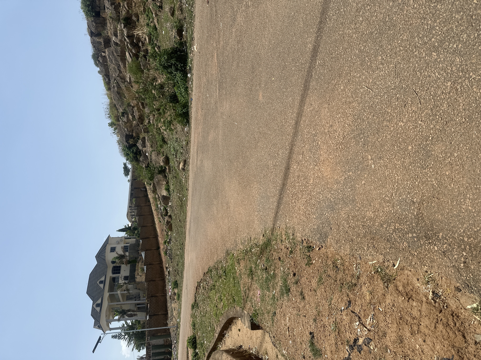 The image shows a paved road winding alongside a rocky hill in Jos, Nigeria. A large house sits behind a brick wall along the road. The environment suggests a somewhat hilly area with sparse vegetation. No people are visible.