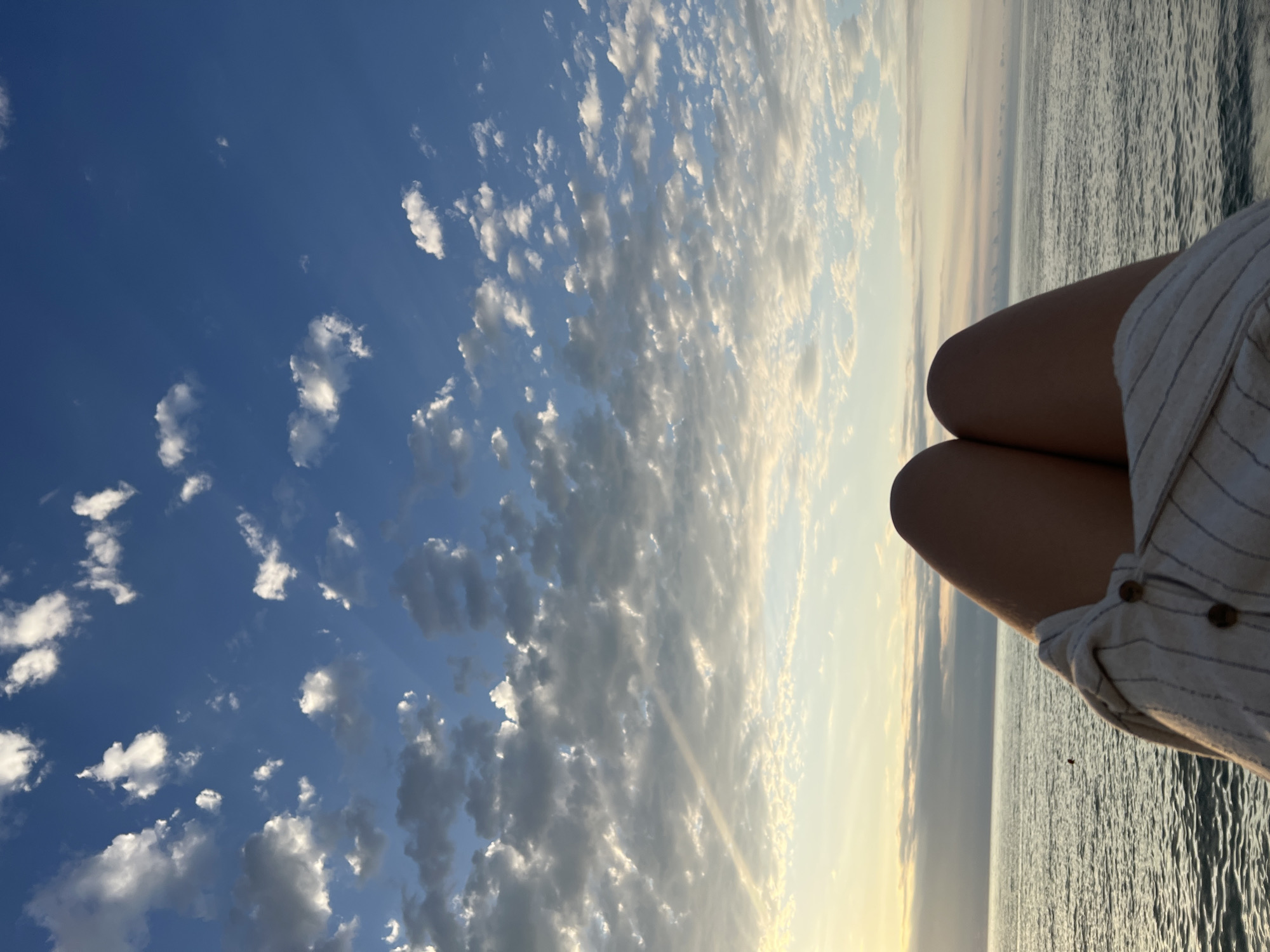 This media file features a close-up shot of a person's legs and torso, clad in a light-colored, striped shirt with visible buttons. The person is seated, with their legs positioned in the foreground. The scene unfolds against a backdrop of a vast, blue sky filled with scattered white and grey clouds. The horizon line, where the sky meets the sea, is visible in the distance. The water appears slightly rippled, suggesting a gentle breeze. The lighting indicates it is either early morning or late afternoon, with the sun casting a warm glow, particularly on the clouds and the horizon. The overall mood is serene and peaceful, with the vastness of the sky and sea evoking a sense of calm. The image was captured in Batumi, Georgia. There is no visible text in the media file.