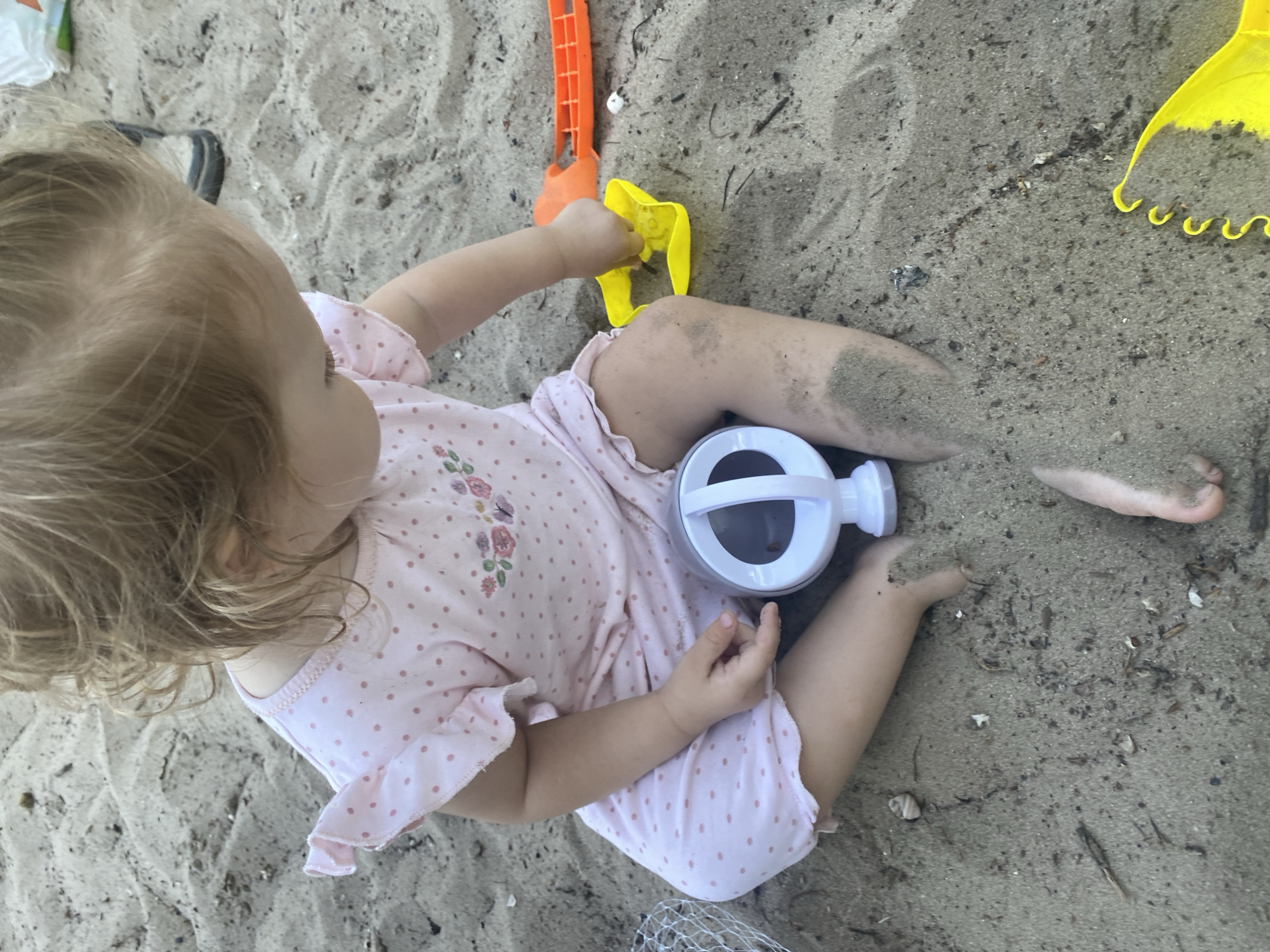 A young child with blond hair sits on sandy ground, wearing a pink polka dot outfit. The child is engaged in playing with sand toys, including a yellow shovel and an orange rake. A small white bucket with a clear plastic lid is also present. The child's legs are partially covered in sand. The sandy environment appears to be outdoors, possibly a beach or playground. The scene is an outdoor setting in Dnipro, Ukraine.