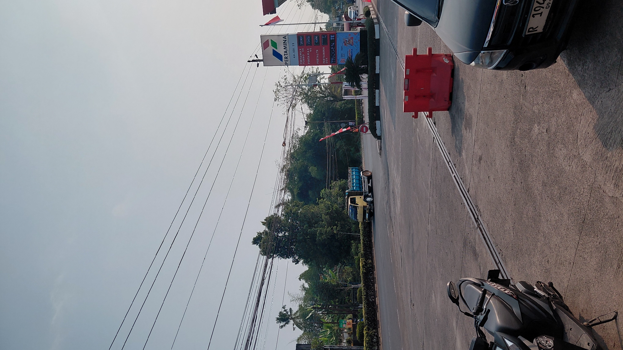 The image depicts a street scene in Karanglewas, Indonesia, likely near a Pertamina gas station. In the foreground, a black scooter is parked on a concrete surface, angled to the left. To the right, the front portion of a black car is visible, with its license plate displaying "R 194". A red traffic barrier is positioned in the middle ground. Across the asphalt road, a yellow truck carrying a load of dark material is driving to the left. Behind the truck, lush green trees and vegetation form a dense backdrop. A tall Pertamina sign, featuring the company logo and fuel prices, stands prominently to the right of the road, with an Indonesian flag attached to its pole. Overhead, numerous power lines crisscross the sky. The sky is a pale blue with a slight haze, indicating daytime.