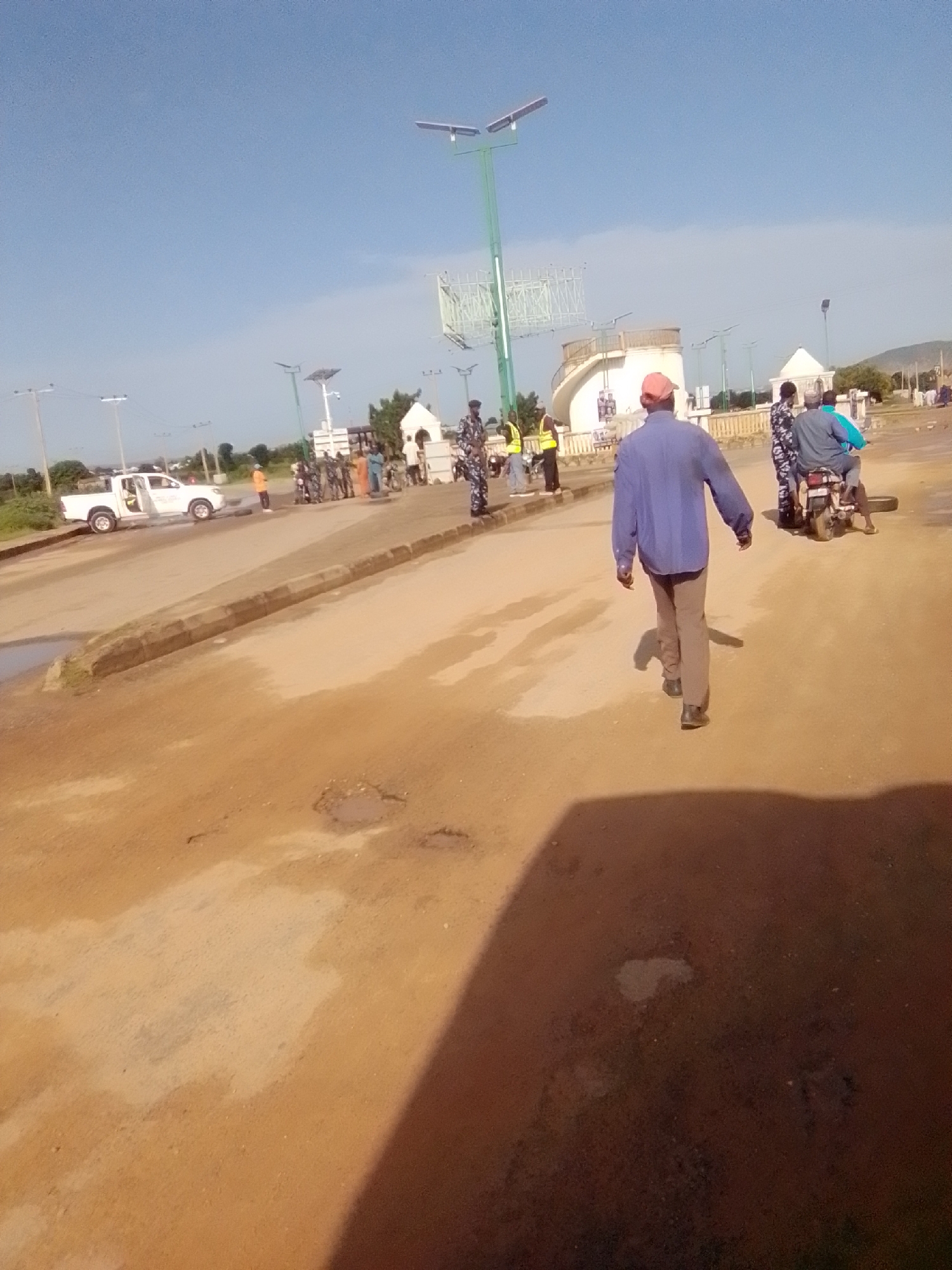 The image captures a scene in the city of Kwadon, Nigeria, on a sunny day with a clear blue sky. The foreground is dominated by a dusty, unpaved road with a few puddles and patches of lighter-colored soil. A large shadow from an unseen object falls across the lower right portion of the frame.
In the middle ground, the road curves to the left and is bordered by a curb. A white pickup truck with its driver's side door open is parked on the left side of the road, facing away from the camera. A person in an orange vest stands near the truck.
Further back, a group of people are gathered, some in what appear to be police uniforms with camouflage patterns. One person is walking away from the camera, wearing a blue long-sleeved shirt and beige pants, with a red cap. Another group of people are standing, and two individuals are seated on a motorcycle, one of whom is wearing a blue shirt.
In the background, there are several streetlights with solar panels, and a white, domed structure with a spiral staircase-like element. Beyond that, trees and buildings are visible, with a gently sloping hill in the far distance. The overall impression is that of a somewhat arid, possibly rural or semi-urban environment.