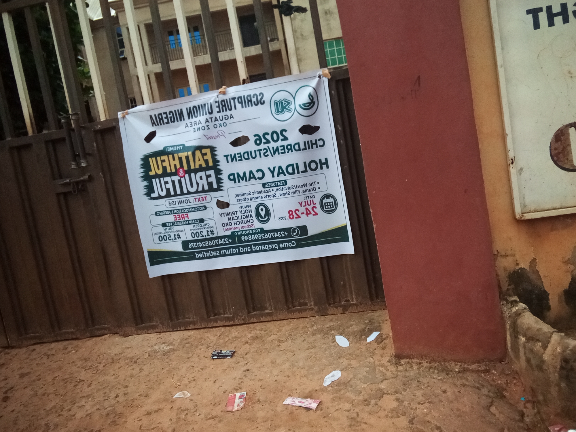 The image captures a banner for a "FAITHFUL & FRUITFUL CHILDREN'S HOLIDAY CAMP" displayed on a metal gate in Oko Town, Nigeria. The banner is white with colorful text and graphics, indicating the event is organized by the Scripture Union of Nigeria, Oko Area, Oko Zone. The camp theme appears to be "Faithful & Fruitful." Details on the banner include dates from July 25-38, which is likely a typo and should be 28, and a fee of #500, suggesting a registration cost. There are also contact phone numbers listed. The background shows a building with windows and a red wall. The ground in front of the gate is dirt with some scattered papers, possibly flyers or wrappers. The overall scene suggests an outdoor community event announcement.