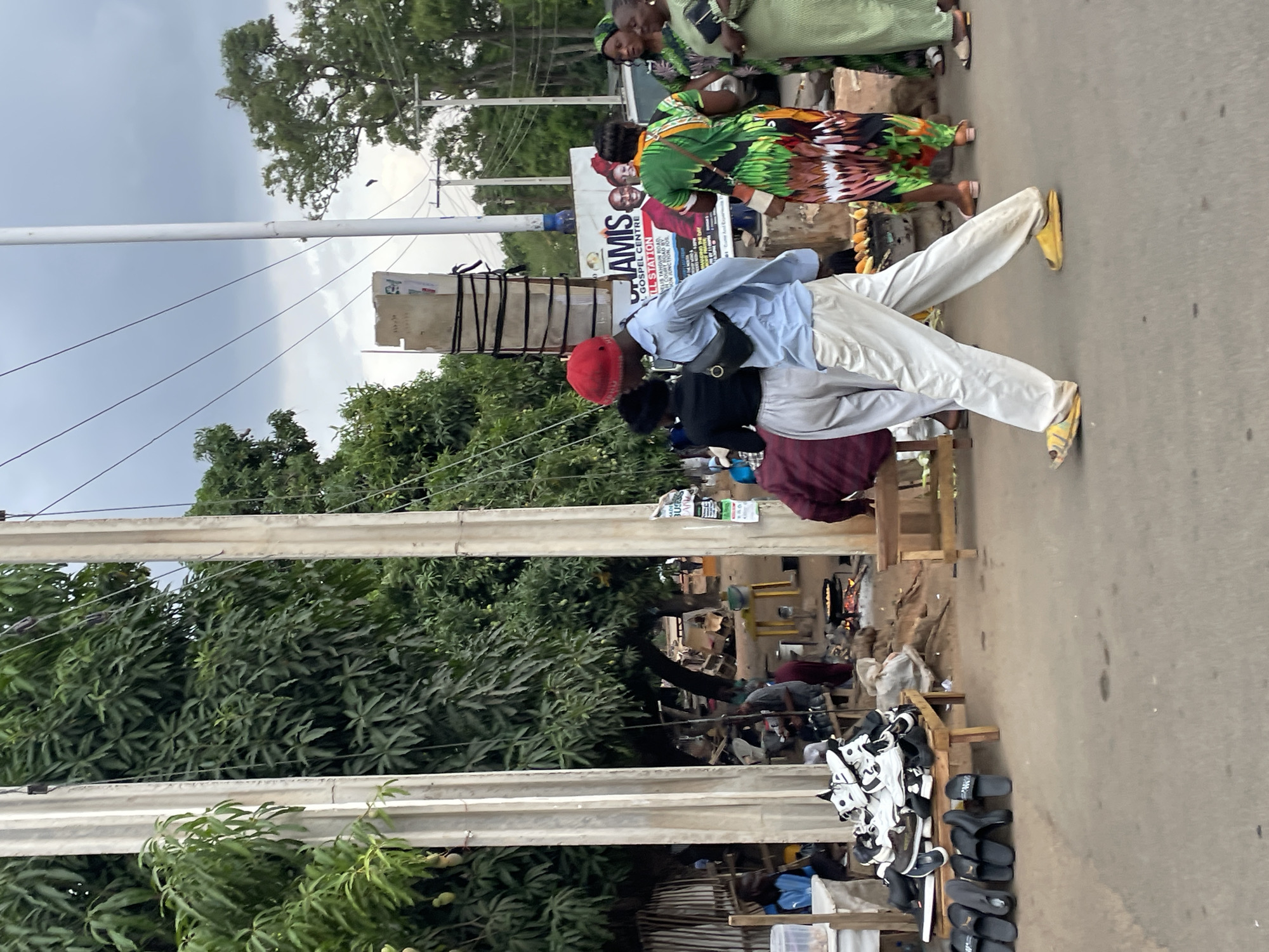 This media file captures a vibrant street scene in Jos, Nigeria, under an overcast sky. The image focuses on a road bustling with pedestrians and market activity.
In the foreground, a man walks across the frame, dressed in a light blue long-sleeved shirt, light-colored pants, and a red cap, with distinct yellow slides/sandals on his feet. He has a dark bag slung across his body. Behind him, another person is partially visible, wearing a dark top. Further along the road, a group of women are seen, one prominently featured in a striking dress with green, brown, and black patterns, and another in a plain light green dress.
To the right side of the road, a series of market stalls are set up. One stall in the immediate foreground displays numerous pairs of shoes, including white sneakers and various black slides and sandals. These items are arranged on wooden display structures. Deeper into the background on the right, more market activity is evident, with people tending to goods and what appears to be a small fire, possibly for cooking or warmth.
The street is flanked by abundant green trees and dense foliage. Utility poles and power lines stretch across the upper portion of the image, silhouetted against the gray, cloudy sky, which suggests a non-sunny time of day. A large billboard is visible behind the foreground figures, displaying images of people. Prominent text on the billboard reads "JAMIS", "GOSPEL CENTRE", and "FELLOWSHIP", with smaller text indicating "TAILIN ROAD" and "JOS", pointing to a local religious establishment. The overall scene depicts a typical day of commerce and movement in a Nigerian urban environment.