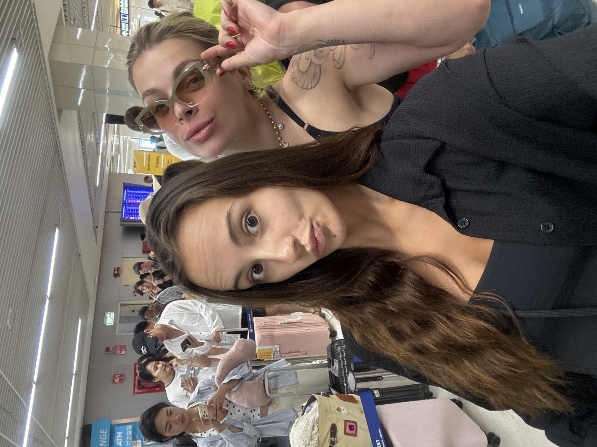 This media file captures two women taking a selfie or group photo in what appears to be an indoor public space, likely an airport or travel hub, specifically in Tambon Mai Khao, Thailand. The image is oriented with the subjects' heads towards the top-left.
In the foreground, a woman with blonde hair, wearing sunglasses and a black top, is positioned towards the upper part of the frame. She has a visible tattoo on her upper arm, red nail polish, and a beaded necklace. Her right hand is raised, holding her sunglasses. Below her, another woman with long dark hair and a dark top is looking towards the camera with a playful pout.
The background shows a bustling environment with numerous other individuals, many appearing to be seated and waiting, possibly with luggage including a pink suitcase and a blue backpack. Overhead, fluorescent strip lighting illuminates the area. Visible cues in the setting include information screens displaying what looks like travel details (though specific content is illegible), and various signs.
Visible text in the scene includes fragmented words like "KET IONA AIR" (likely part of an airport name), "NGE ATM" (suggesting Exchange and ATM services), "EXIT," "ช่องถามพิเศษ" (Thai for "Special Inquiry Channel" or "Priority Lane"), "Priority," "Seat," and the brand name "QUECHUA" on a bag. There are also less clear texts such as "AN DIO," "AOT/PRAET," and "wheren." The atmosphere suggests a travel setting, with people waiting and engaging with their phones. The emotions conveyed by the main subjects are casual and playful.
