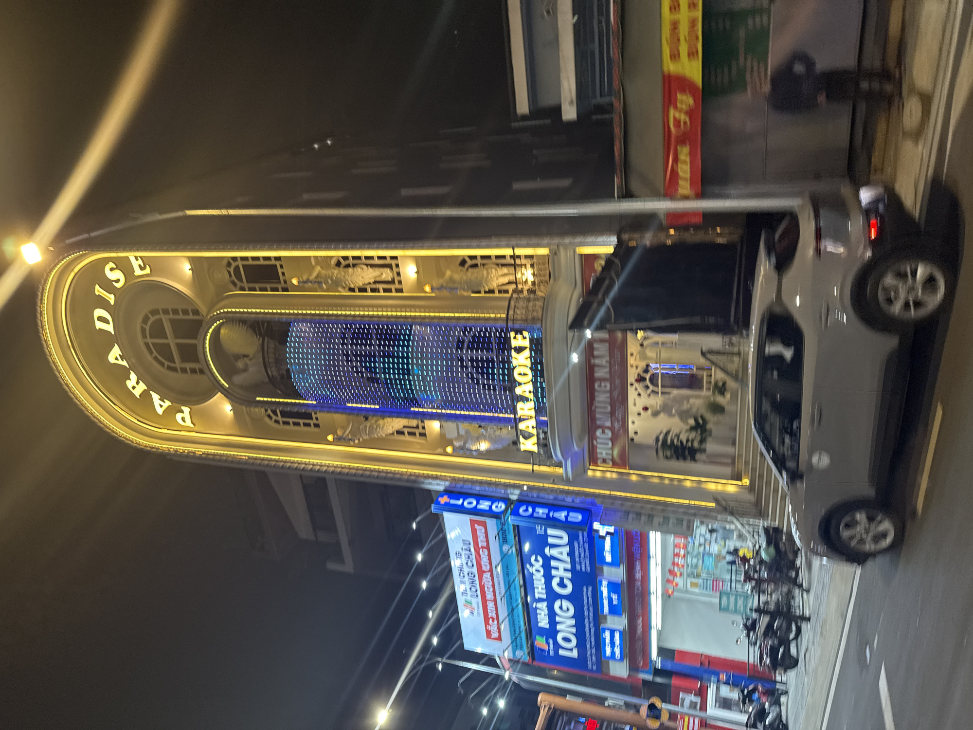 The image captures a vibrant night scene on a street in Cam Ly - Đà Lạt, Vietnam. The main subject is a large, brightly lit building housing "PARADISE KARAOKE." The building's facade is adorned with yellow illuminated trim and features a prominent blue LED screen with shifting patterns.
On the street level, a white car is parked, and several motorcycles are lined up, suggesting a commercial area. Streetlights cast bright beams, cutting through the dark night sky. Other businesses are visible, including "NHÀ THUỐC LONG CHÂU" (Long Chau Pharmacy/Drugstore), indicated by several prominent blue and white signs. One sign for Long Chau mentions "VẮC XIN NGƯỜA UNG THƯ" (Cancer Vaccine) and a phone number starting "1800-E". Another sign on the karaoke building reads "CHÚC MỪNG NĂM" (likely "Happy New Year"). A person is partially visible near the parked car. The overall impression is a lively urban environment after dark.