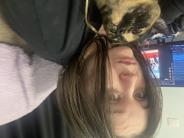 The image depicts an indoor scene featuring a young adult female and a calico cat. The specific location within Pushchino, Russia, is not discernible from the visual evidence.
The female subject has dark, medium-length hair, appearing somewhat damp, parted on the side and falling forward. She possesses light-colored eyes and a neutral expression, gazing directly at the viewer. She is wearing a dark, long-sleeved garment, with her left shoulder and upper arm visible.
A calico cat with black, orange, and white fur patterns is positioned in the lower-left portion of the frame, resting against the female subject's torso. The cat's head is tilted upwards, and its eyes are partially closed.
In the background, a computer monitor is visible to the left of the female subject, displaying a screen that includes what appears to be a YouTube interface. A video featuring individuals on a red carpet is displayed on the monitor. To the left of the monitor, a white electrical outlet or switch is mounted on a light-colored wall. To the right of the female subject, a light purple, textured fabric object, possibly a blanket or cushion, is present against a dark background. The scene is illuminated by interior lighting.