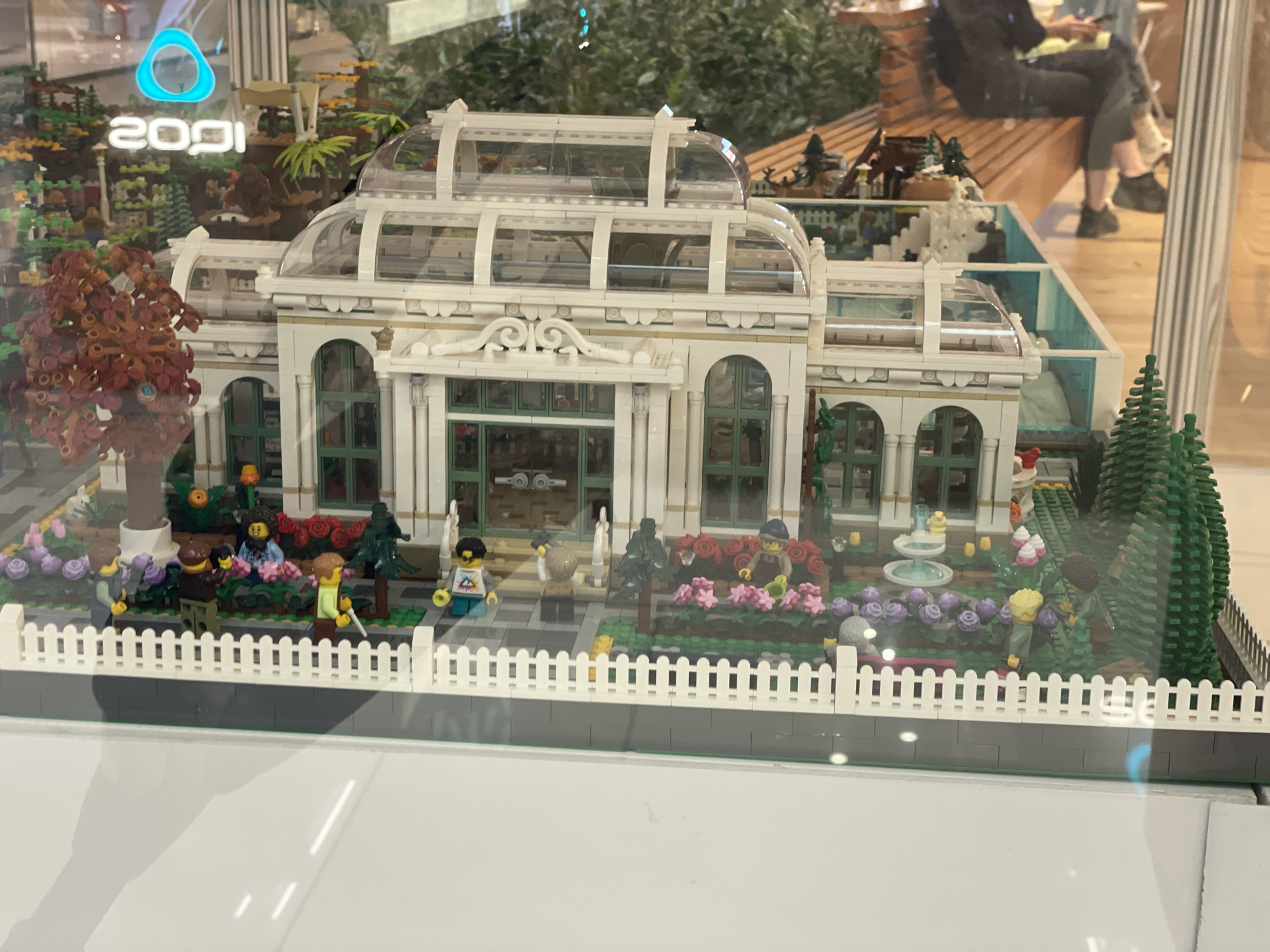 The image shows a detailed Lego model of a Victorian-style greenhouse, complete with a garden and several Lego figures. The greenhouse is white with clear domed roofs and green accents. In front of the greenhouse, there is a manicured garden with colorful flowers, including pink and purple roses, as well as trees. Several Lego figures are present in the garden, some appearing to be tending to the plants, while others stand by. A white picket fence surrounds the garden area. In the background, through the glass, there are blurred figures of people and some retail displays with logos such as "2091" and a blue triangular logo. The overall scene depicts a meticulously constructed miniature garden and greenhouse. The presence of the "2091" logo suggests this display may be located in a mall or store within Sofia, Bulgaria, as the company "2091" is known for its technology and retail presence in the city.