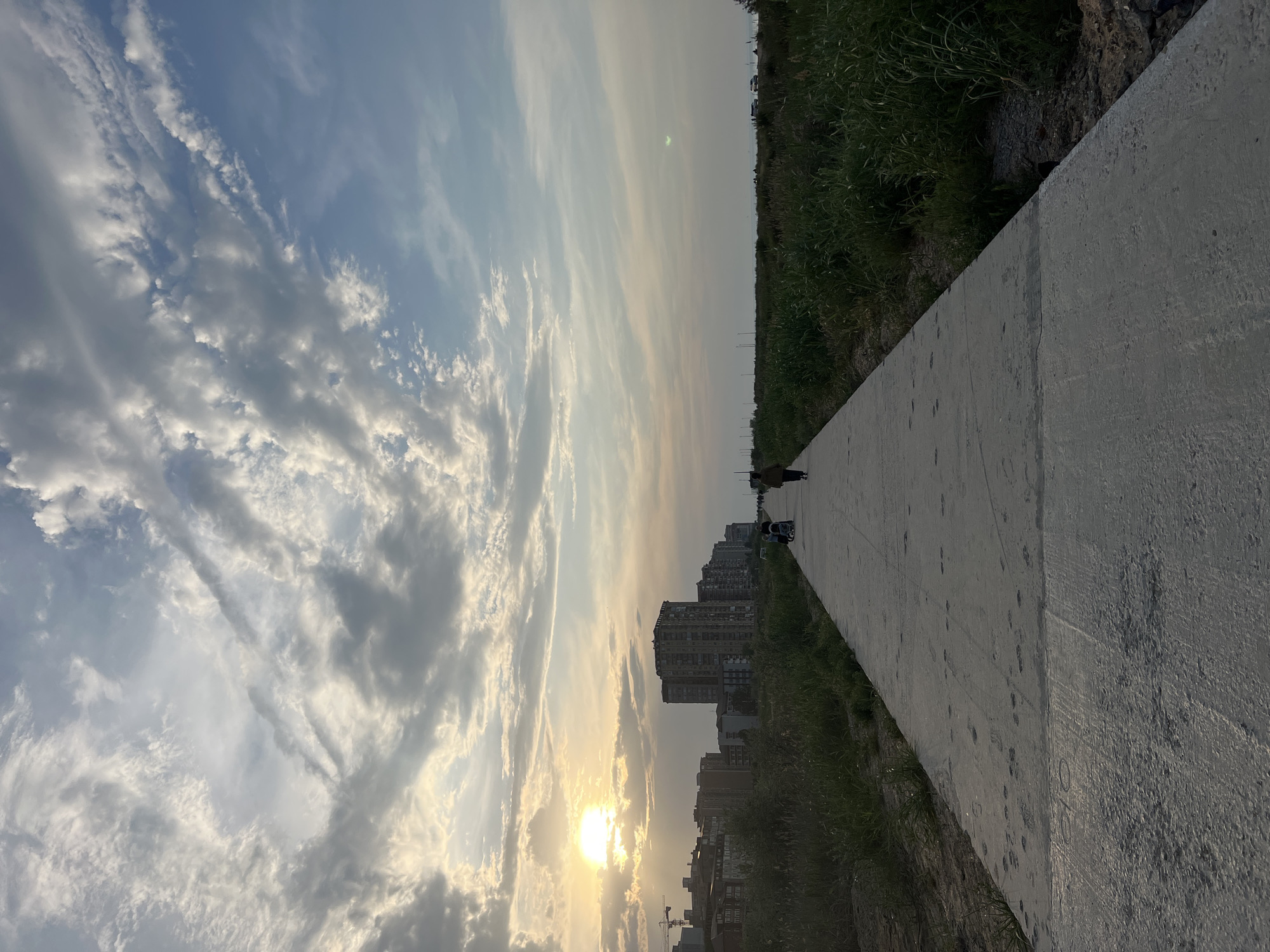 The image depicts a sidewalk stretching into the distance under a cloudy sky with the sun near the horizon, suggesting it may be sunset. Buildings are visible along the horizon, adjacent to the sidewalk, and there is vegetation on the other side of the sidewalk. The location appears to be urban. People are also seen on the sidewalk.